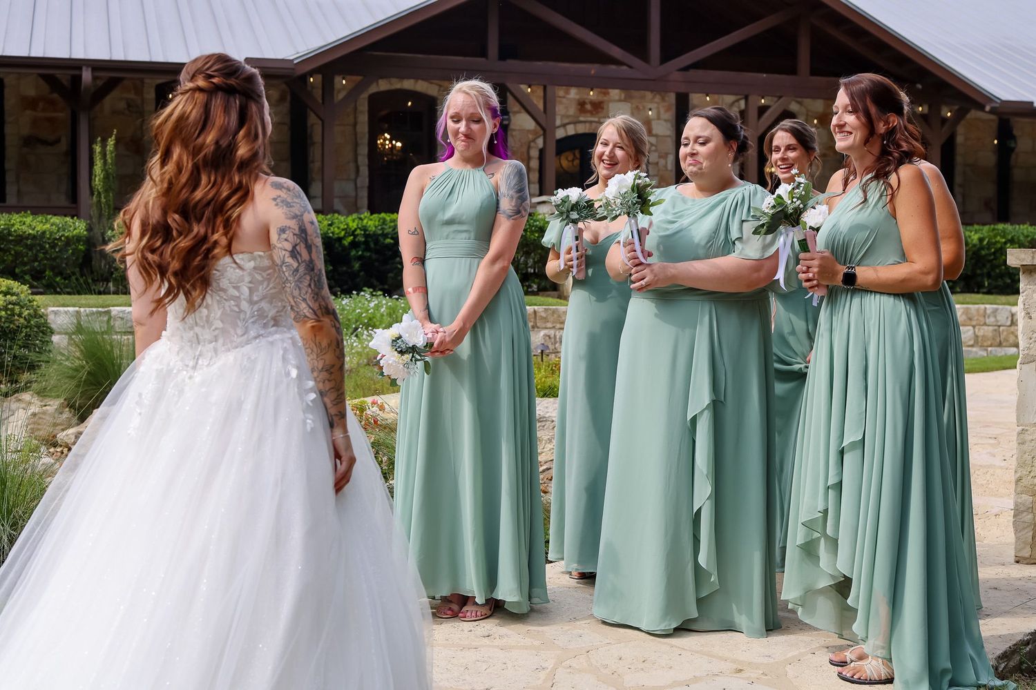 Bride in lace gown reveals herself to emotional bridesmaids in sage green dresses outdoors.