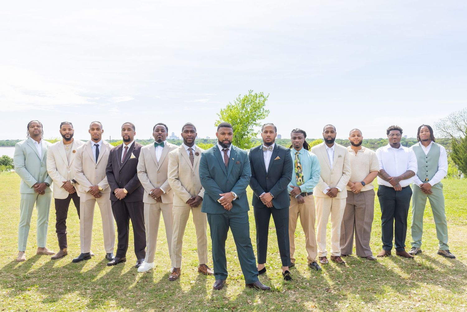 Groom and groomsmen in various colored suits posing outdoors on green lawn for wedding party photo.