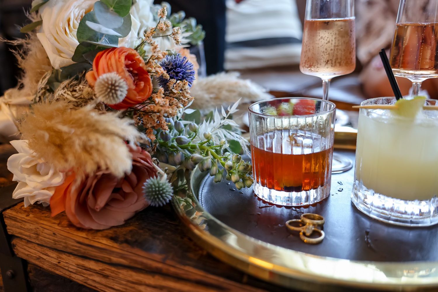 Wedding cocktail hour setup featuring colorful floral bouquet beside amber and white craft cocktails on a gold tray.