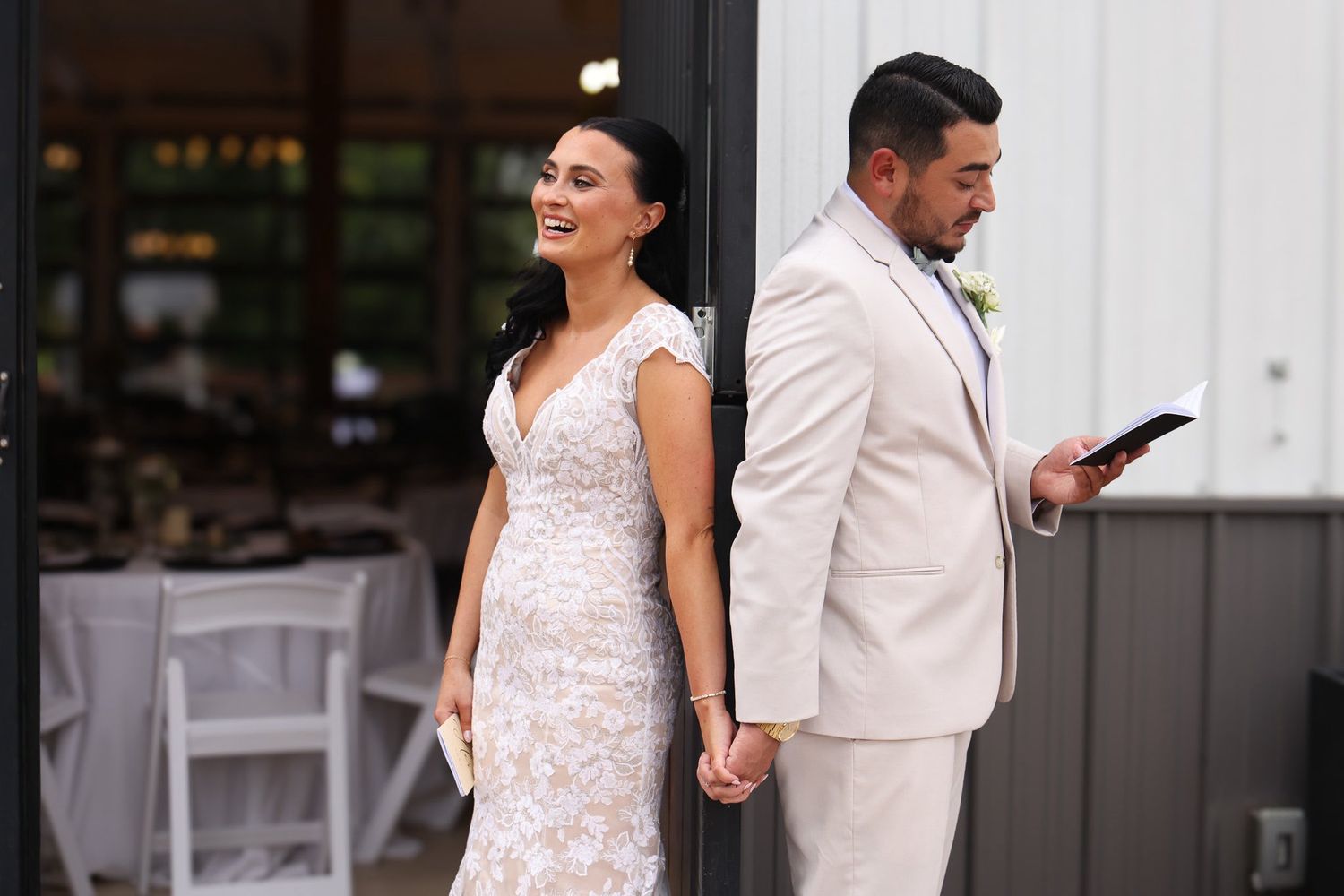 Bride in lace wedding gown and groom in tan suit share a first look moment holding hands.