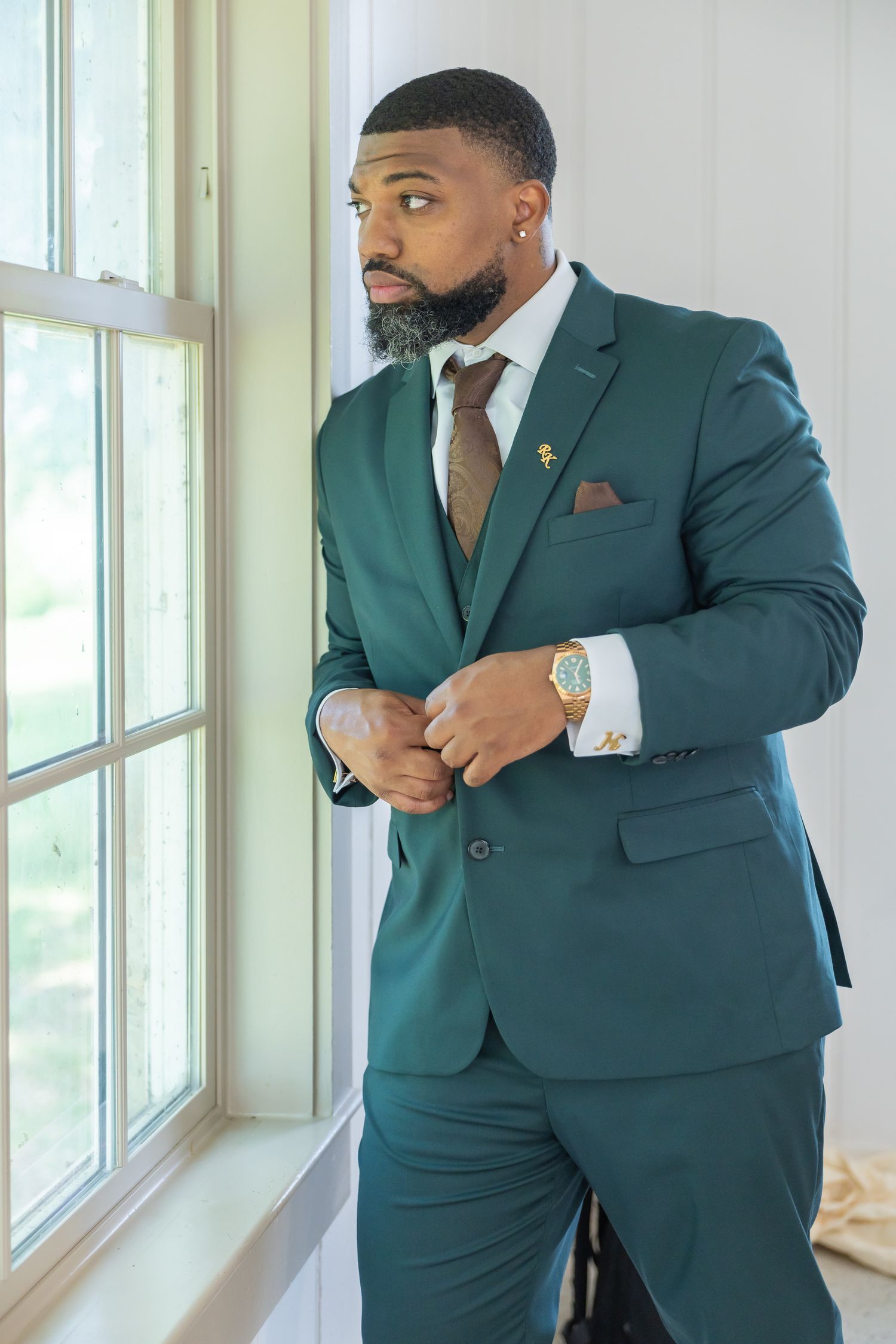 Handsome groom in emerald green three-piece suit buttoning jacket while standing by window.