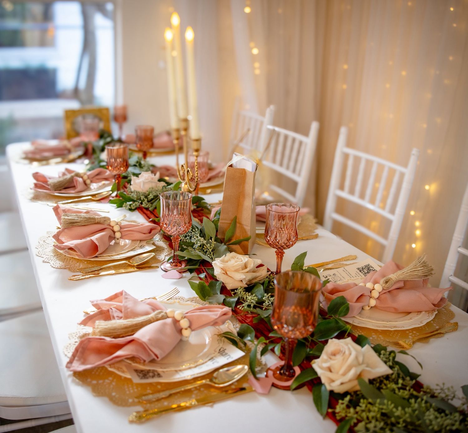 Romantic wedding reception table with pink napkins, gold chargers, candles, roses, and fairy light backdrop.