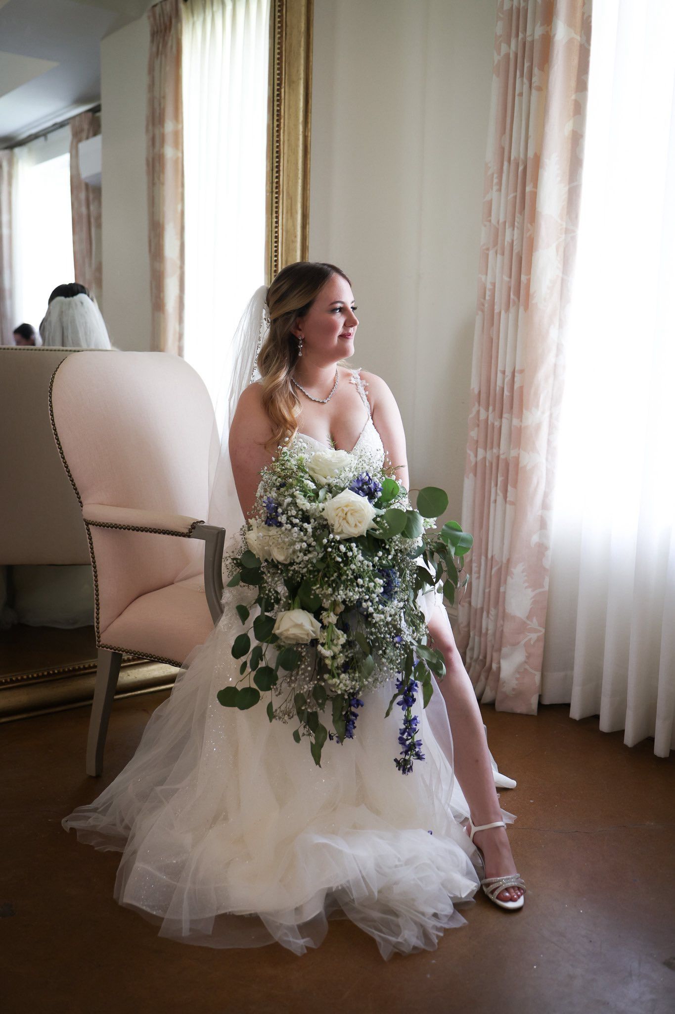 Bride in a tulle wedding gown holding a lush white and blue cascading bouquet while seated by a window.