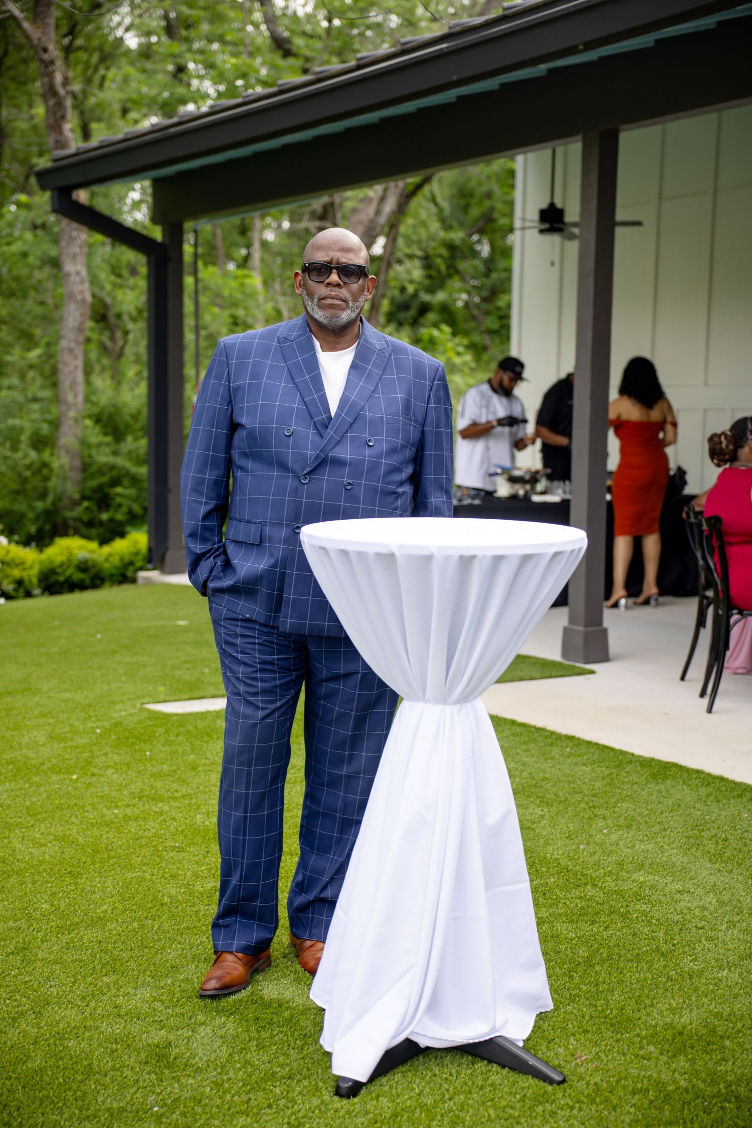 Man in blue plaid suit standing beside a cocktail table at an outdoor garden party event.