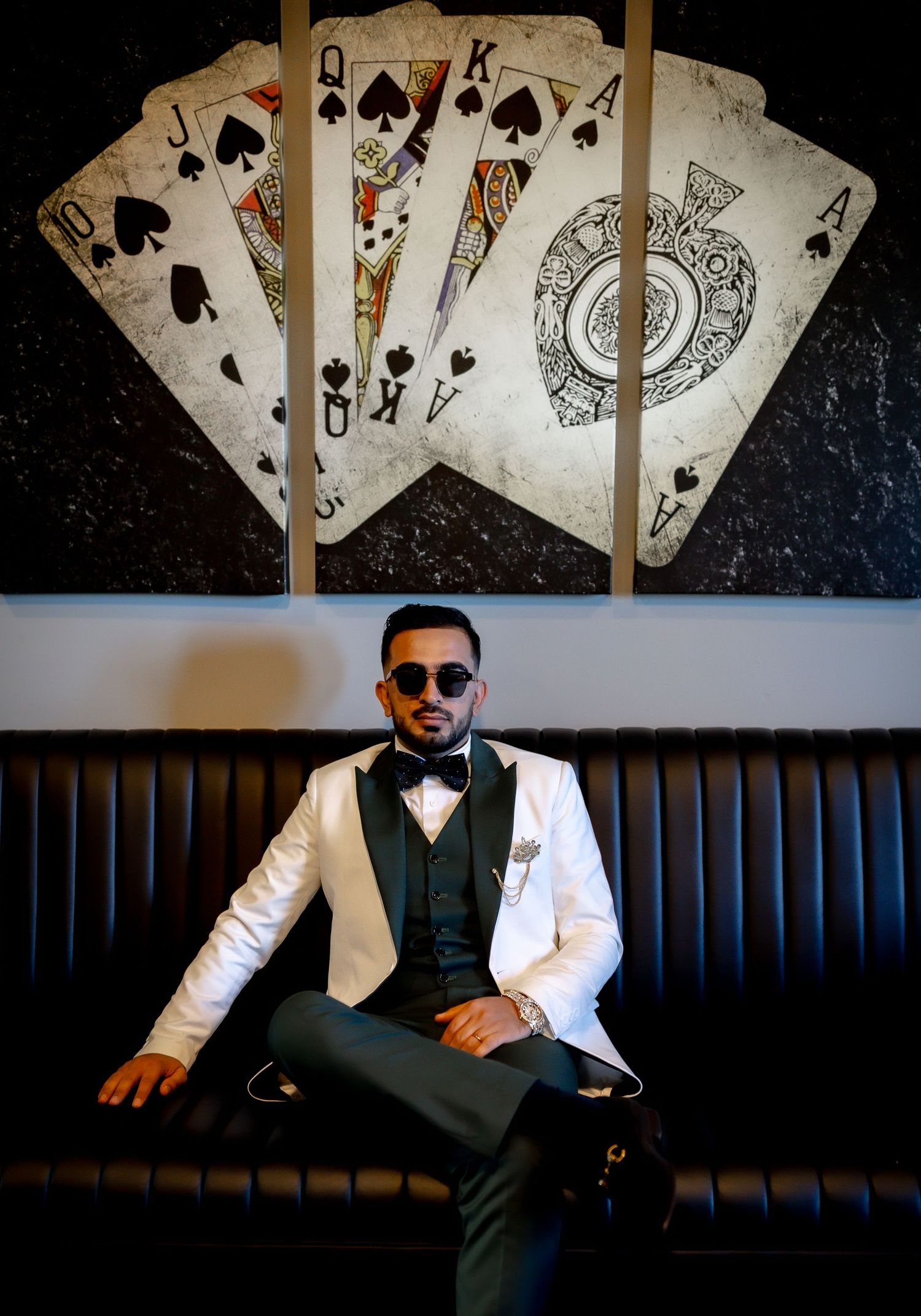 A stylish man in a white tuxedo jacket sits confidently on a leather sofa beneath a large playing cards wall art.