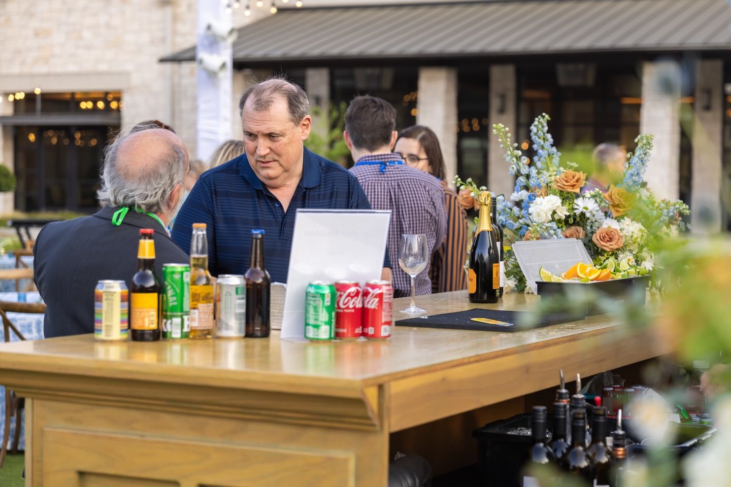 A man stands at an outdoor wooden bar surrounded by beverages and floral arrangements.