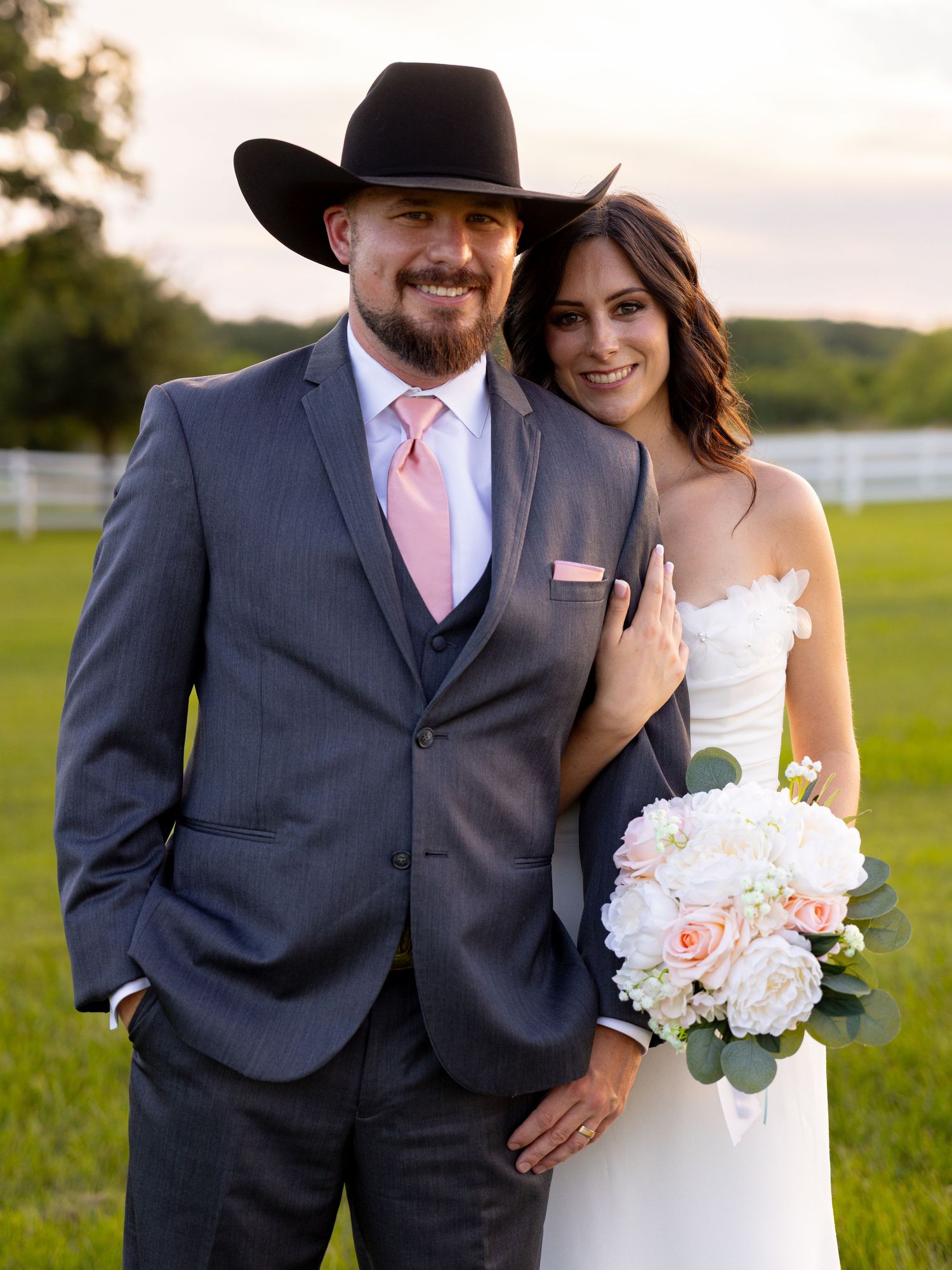 Smiling couple poses together on their wedding day, groom in gray suit and cowboy hat, bride holding pink bouquet.