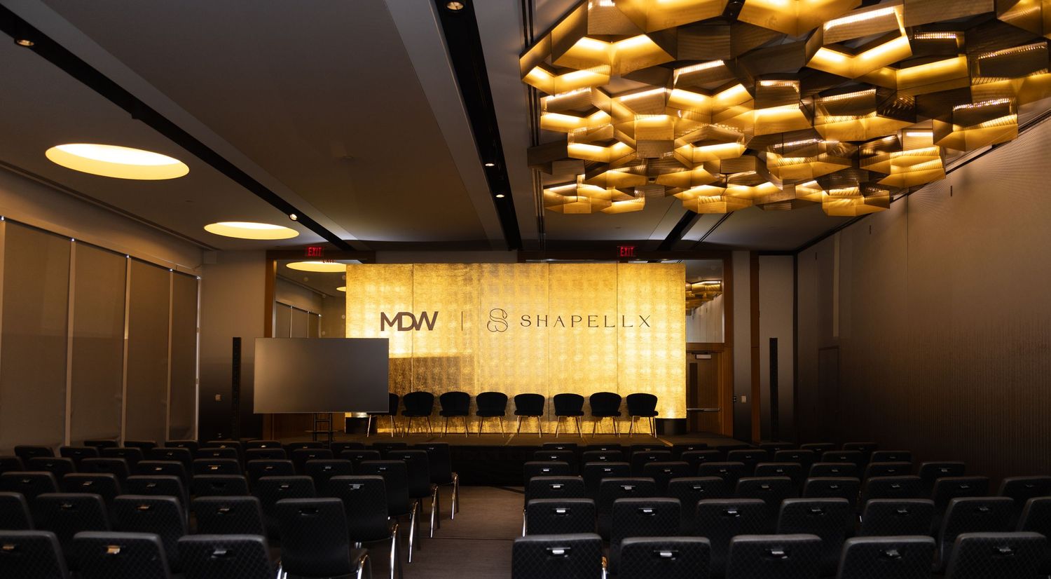 Modern conference hall with golden honeycomb ceiling lights, rows of seats, and illuminated MDW Chapelle stage backdrop.