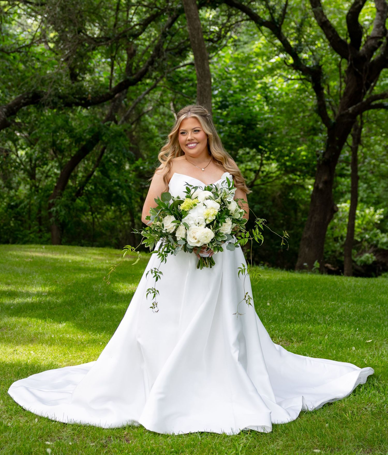 Elegant bride in a strapless white ball gown smiles while holding a white and green floral bouquet outdoors.