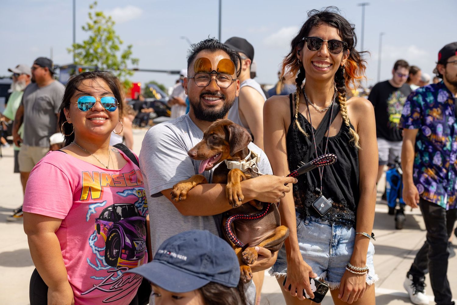 Three friends smile outdoors at a sunny event, with a man holding a small brown dachshund dog between two women.