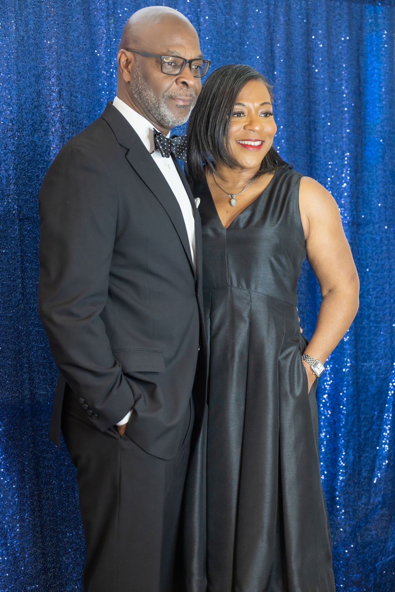 A couple dressed in black formal wear poses together in front of a blue sequin backdrop.