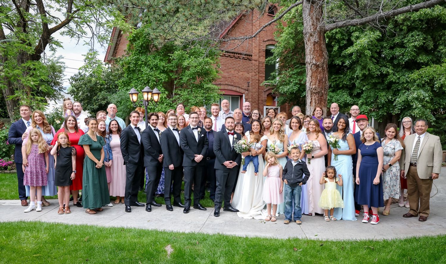 Large wedding party group portrait outdoors on green lawn beside historic red brick building.