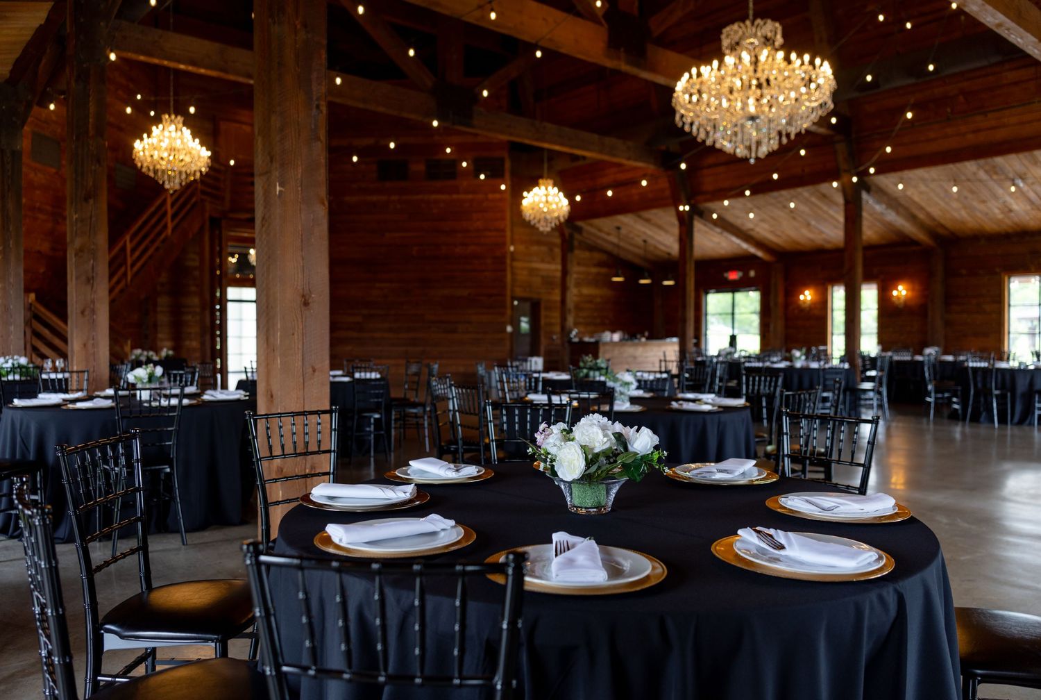 Rustic barn wedding reception venue with black tablecloths, gold chargers, and crystal chandeliers overhead.