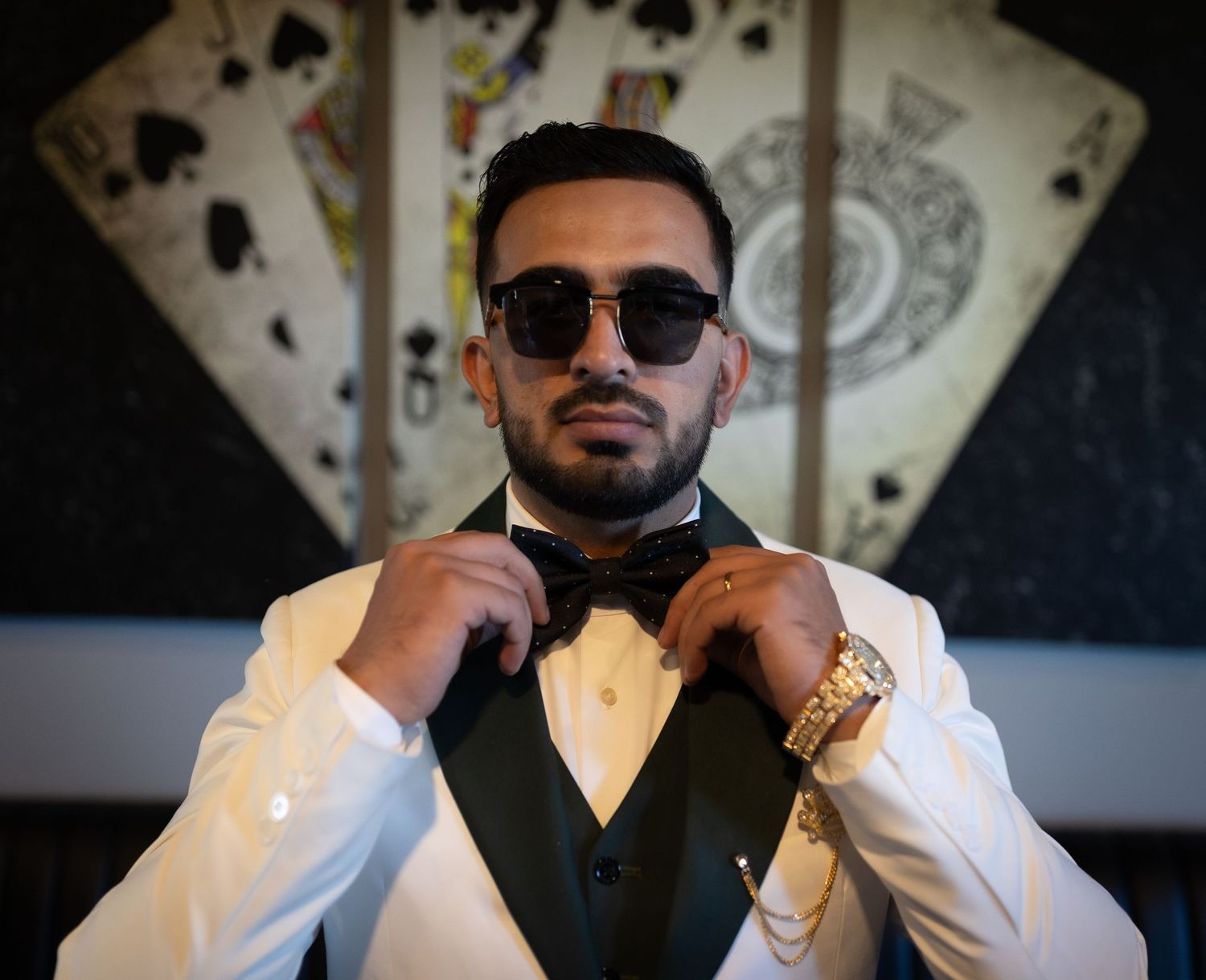 Stylish man in white tuxedo adjusting bow tie, wearing gold watch and sunglasses, with playing cards backdrop.