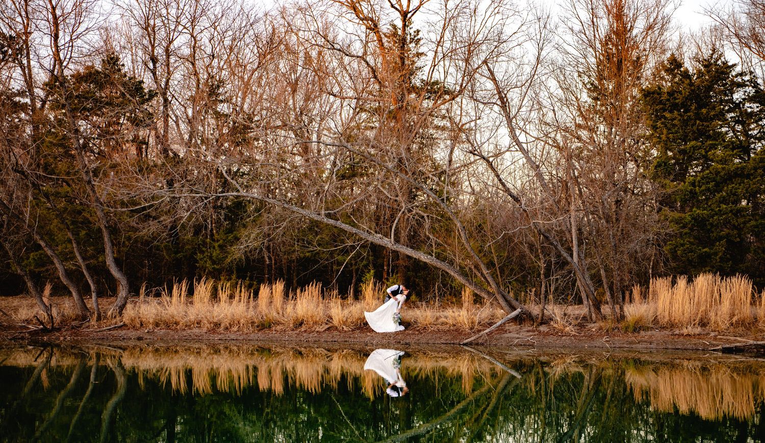 Bride in white gown reflected in calm pond surrounded by autumn trees at golden hour.