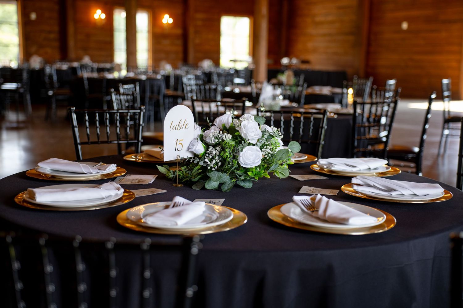 Elegant wedding reception table set with black linens, gold chargers, white floral centerpiece, and chiavari chairs.