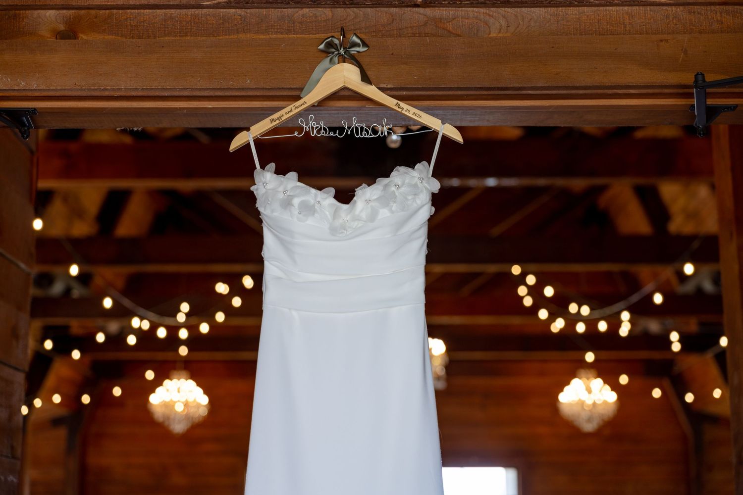 Elegant white wedding dress displayed on a personalized hanger inside a rustic barn with warm string lights.