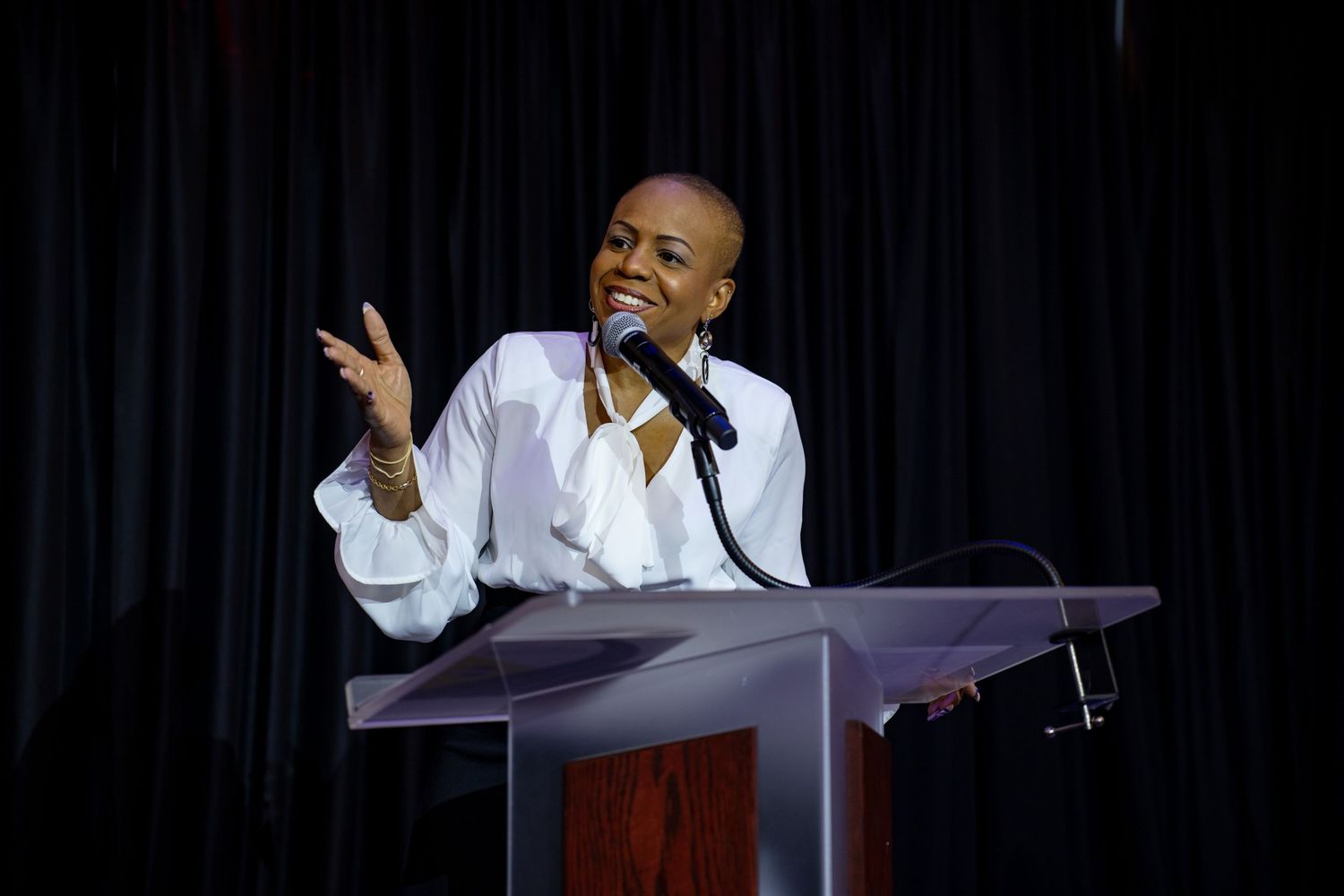 A woman with a shaved head speaks passionately at a podium wearing a white blouse against a dark backdrop.