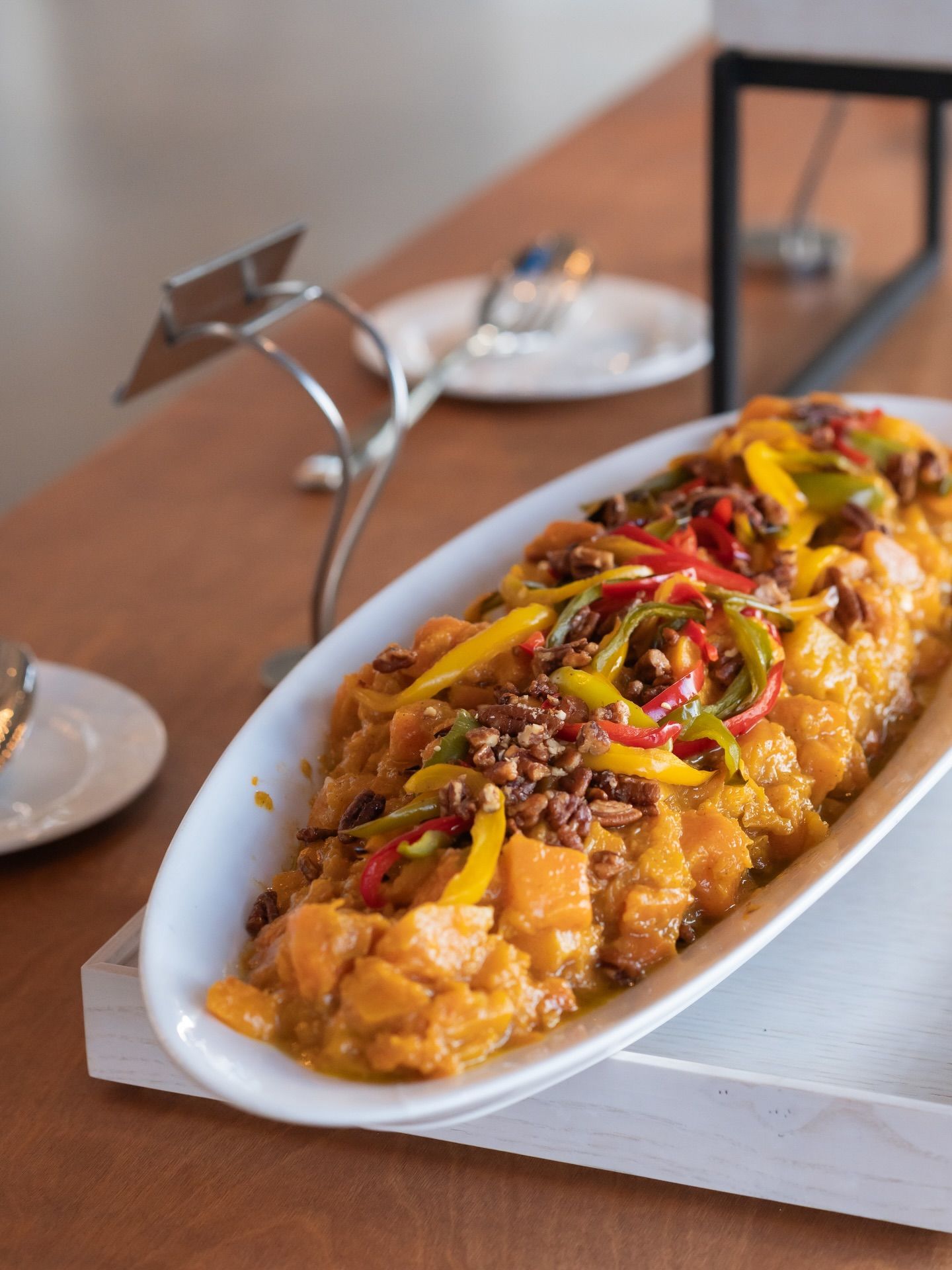 Elegant oblong white dish of seasoned chicken topped with colorful peppers and nuts on a wooden table.