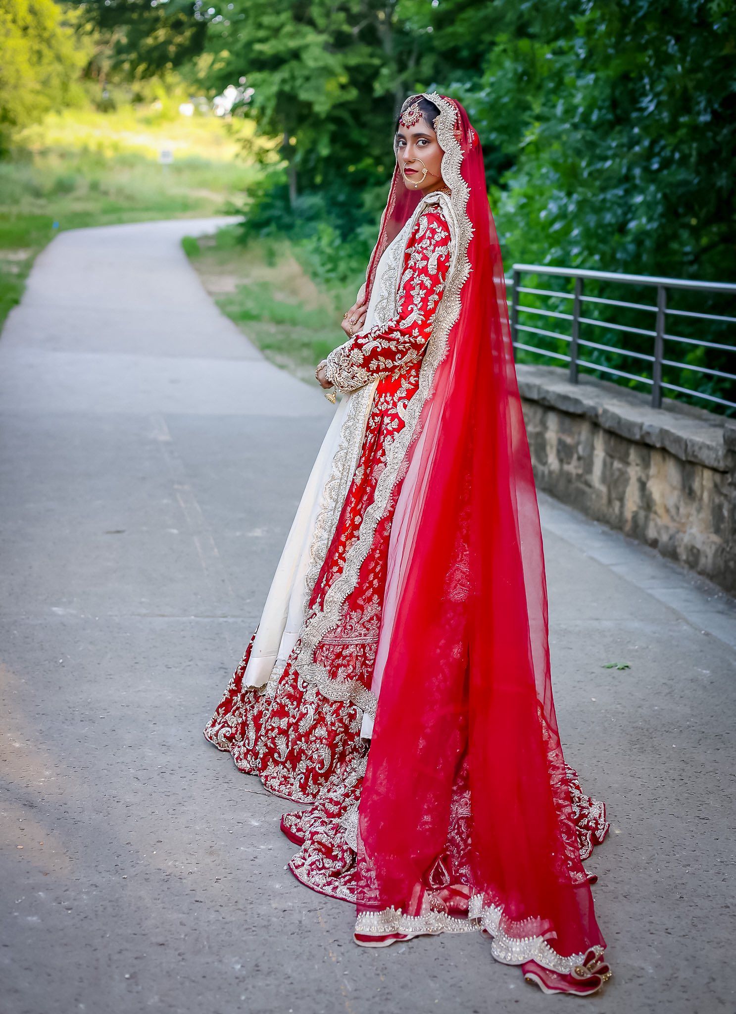 A bride in a stunning red and white traditional South Asian bridal lehenga with a flowing red dupatta on a path.