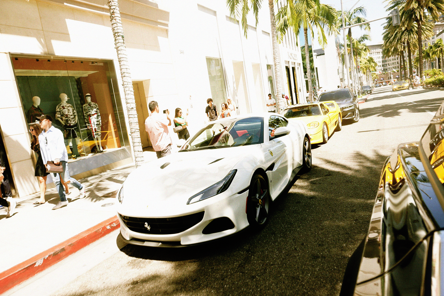 White Ferrari convertible parked on an upscale Rodeo Drive street with shoppers and palm trees.