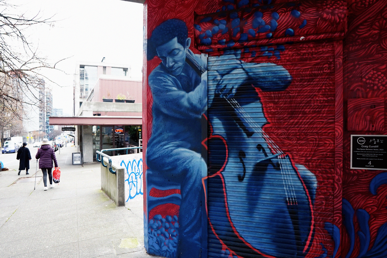 Large colorful graffiti mural depicting a man playing cello painted in red and blue on city wall.