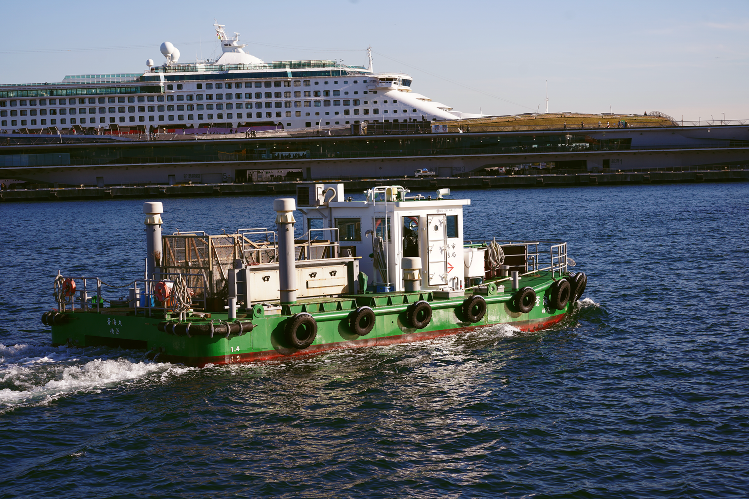 Green tugboat navigating a harbor with a large white cruise ship in the background.
