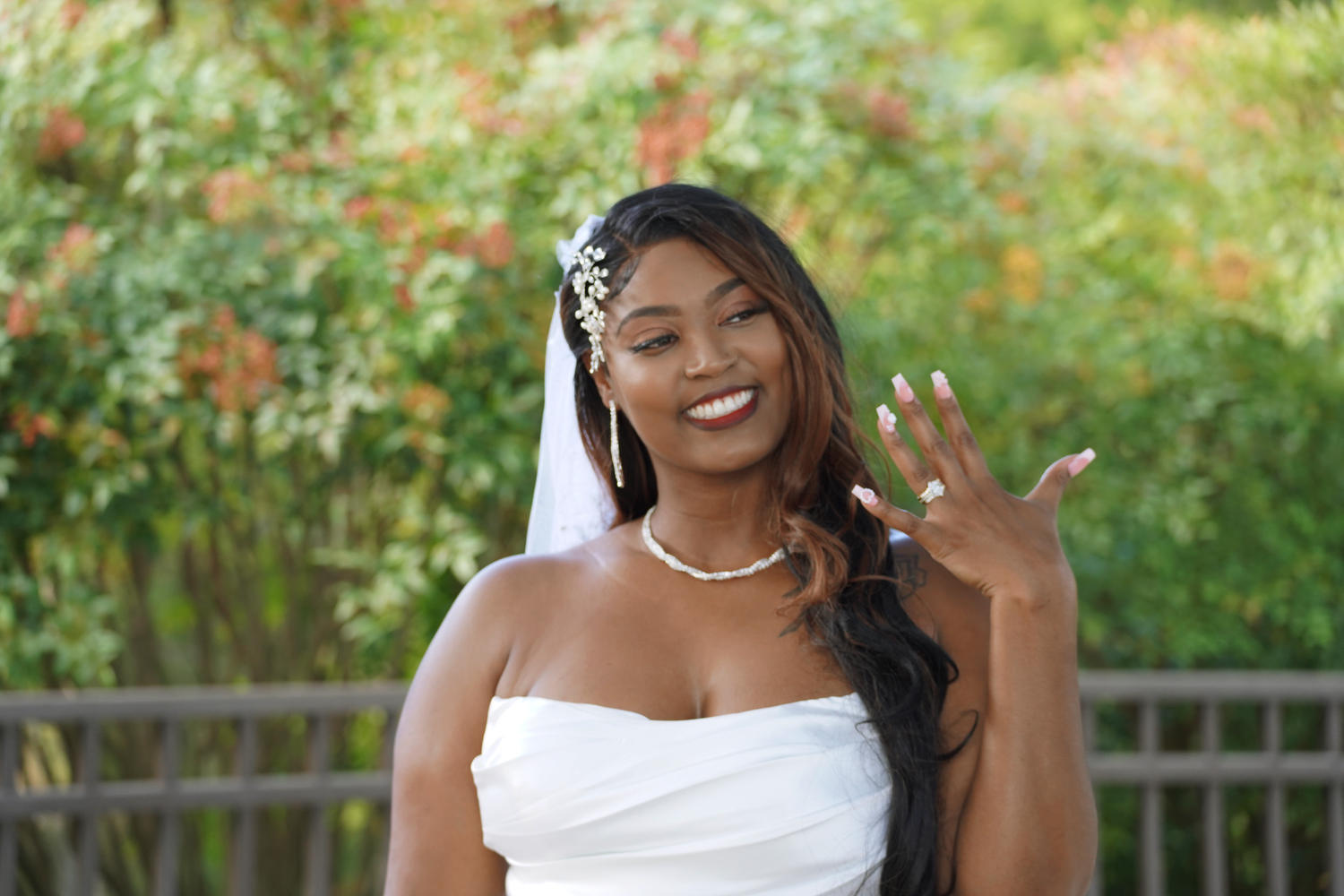 Smiling Black bride in white strapless gown showing wedding ring with floral veil and pearl necklace.