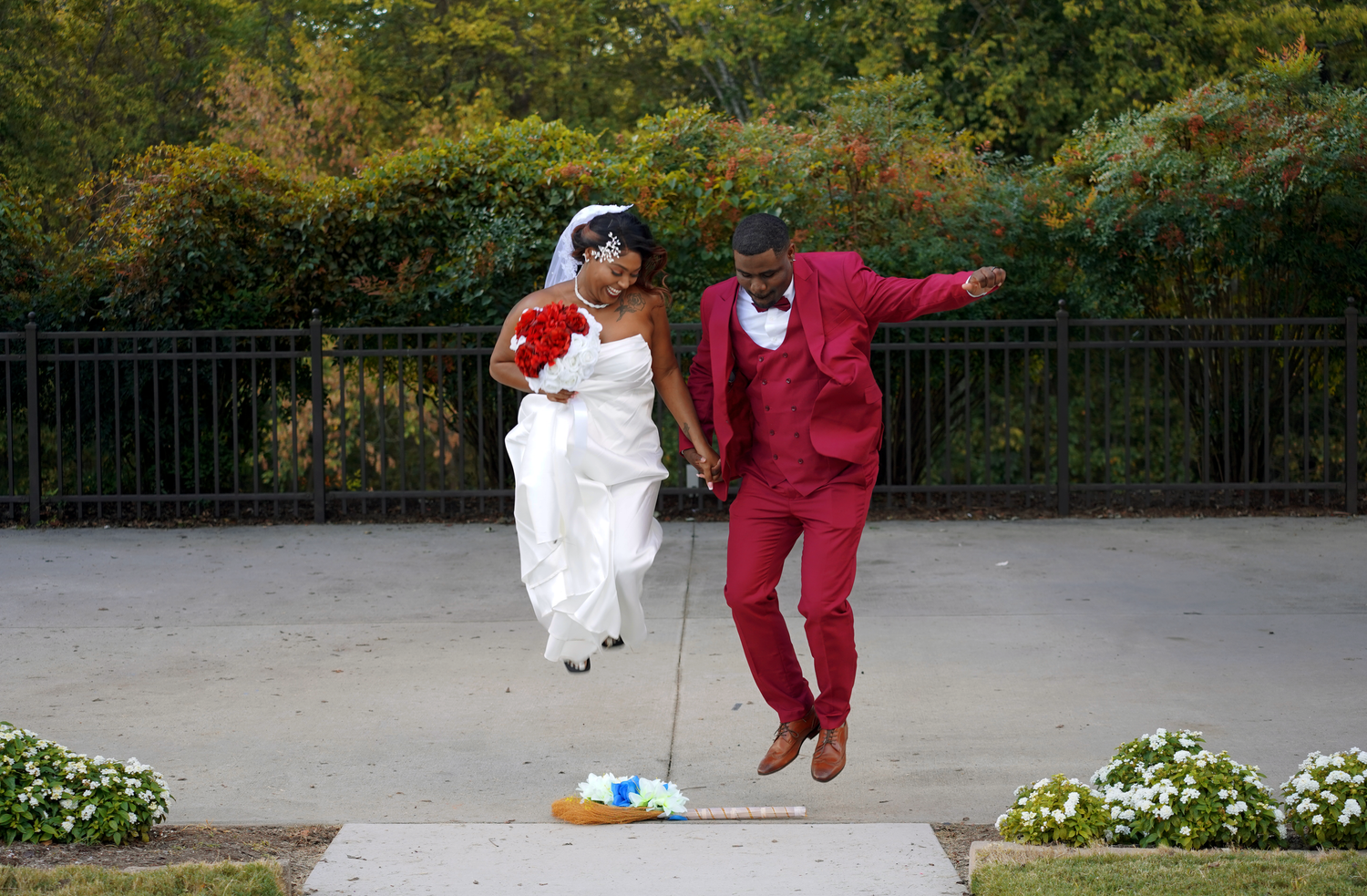 Joyful newlywed couple jumping together outdoors, bride in white gown and groom in burgundy suit.