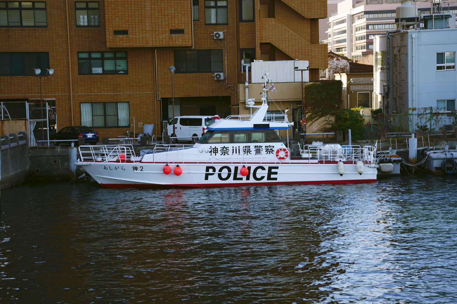 A white Japanese police boat docked in an urban waterway with POLICE written in bold red letters on its hull.