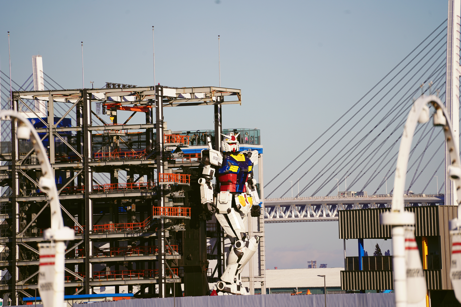 Life-size Gundam robot statue stands before industrial scaffolding with a suspension bridge behind.