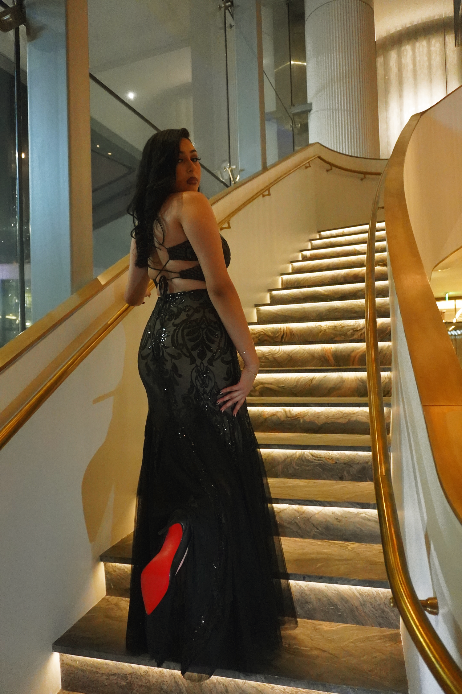 Woman in elegant black mermaid gown with red-sole heels on golden staircase.