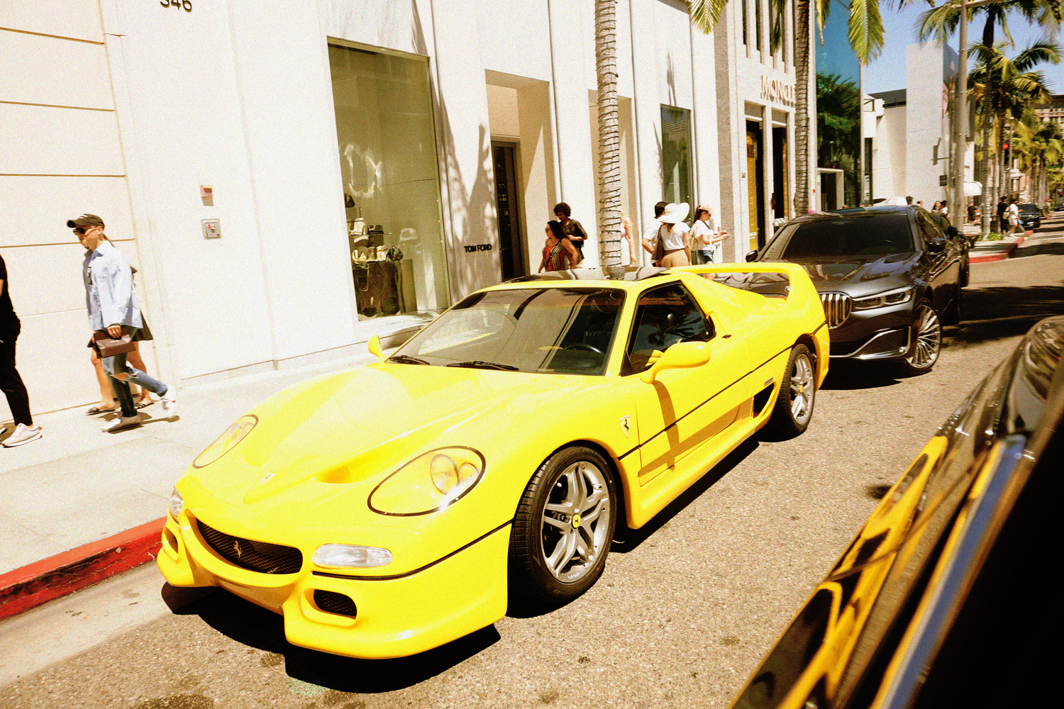 Bright yellow Ferrari F50 supercar parked on a luxury shopping street lined with palm trees.