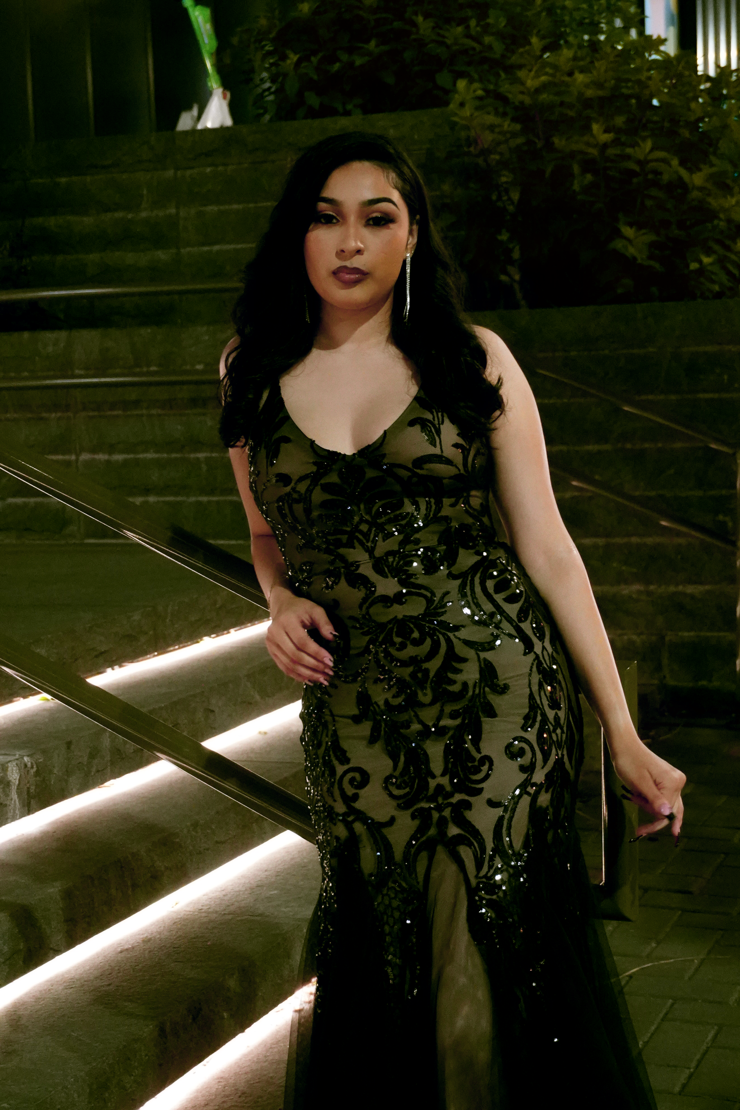 Woman in elegant black sequin mermaid gown posing on illuminated staircase at night.