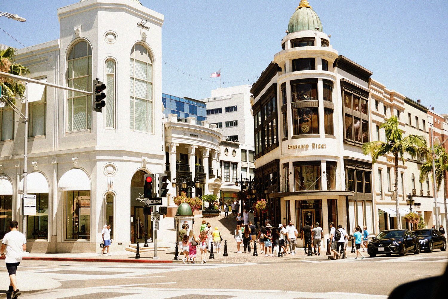 Busy Rodeo Drive intersection in Beverly Hills with luxury shops, pedestrians, and palm trees on a sunny day.