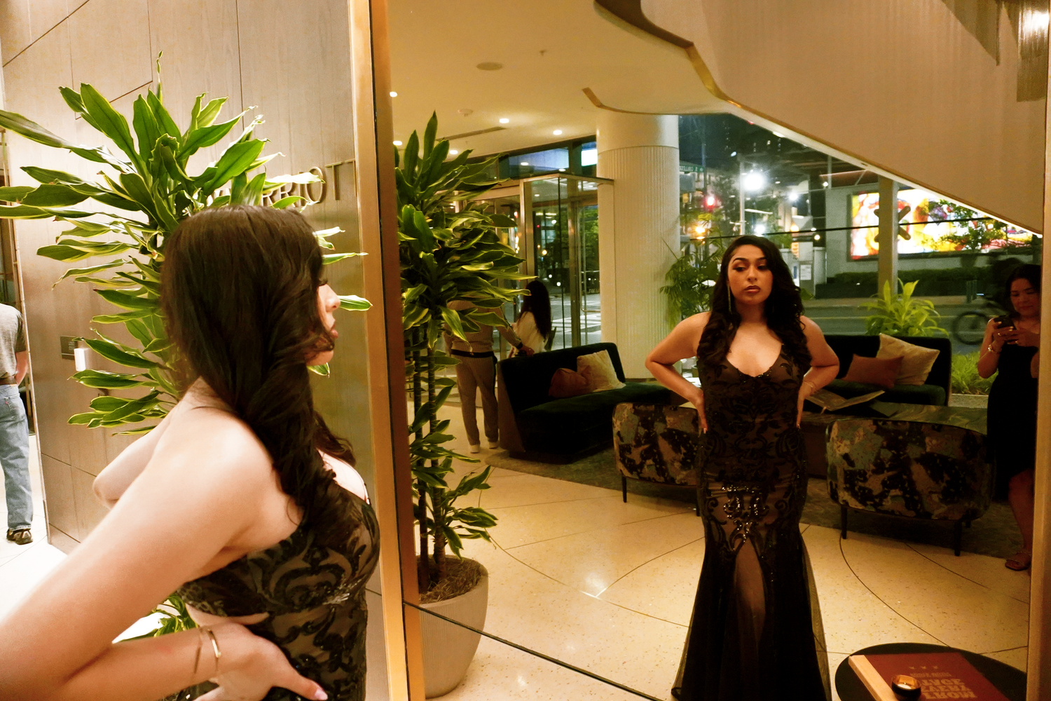 Woman in black lace gown posing in front of a mirror in a luxury lobby.