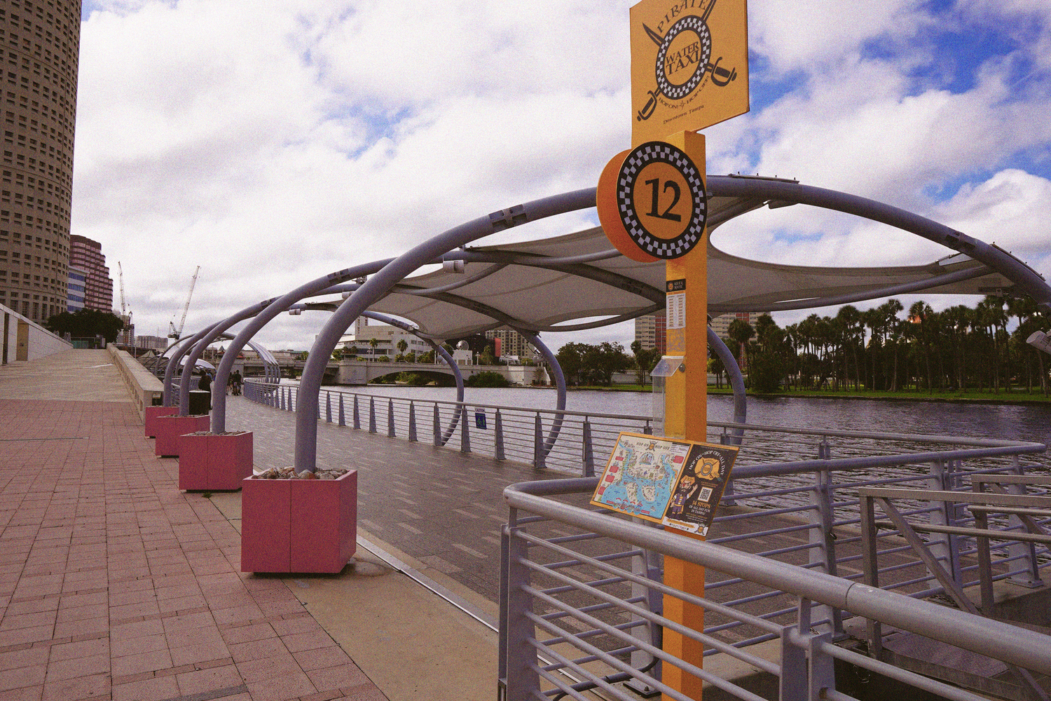 A covered pedestrian waterfront walkway with arched canopy structures along the Hillsborough River in Tampa.
