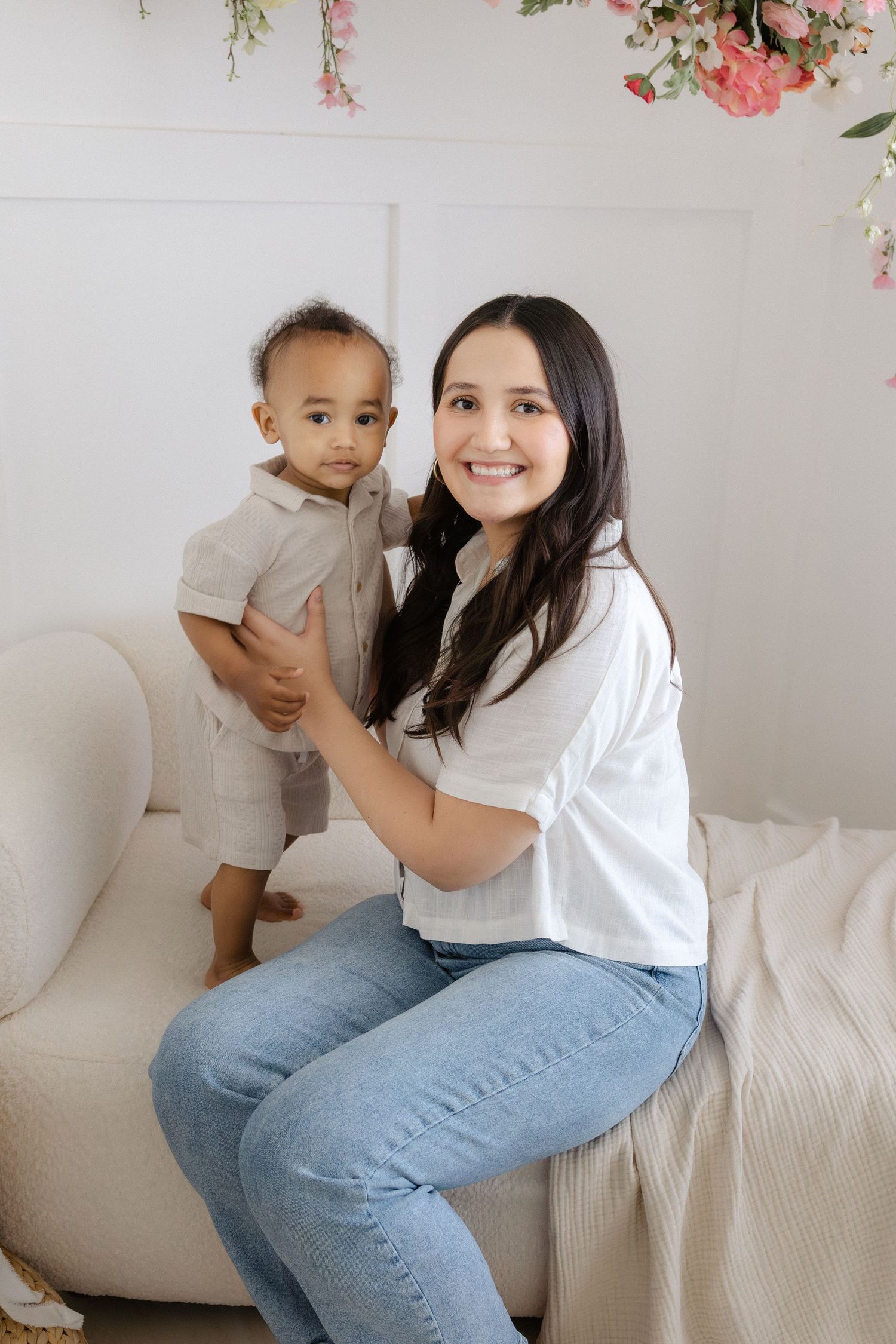 A smiling woman in white shirt and jeans sits on a couch holding a baby boy in a bright floral studio.