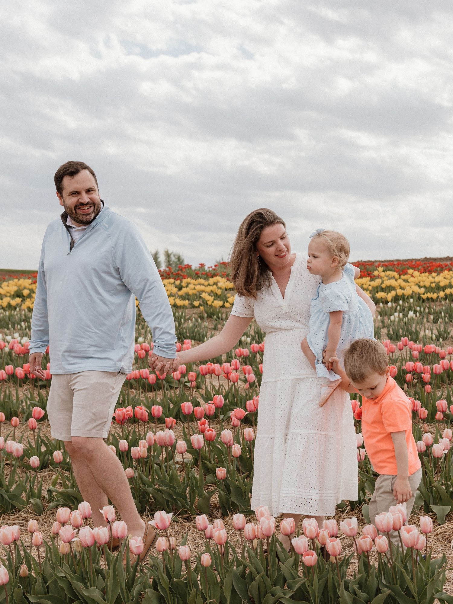 Family of four posing in a vibrant tulip field with pink, yellow, and red blooms under a cloudy sky.