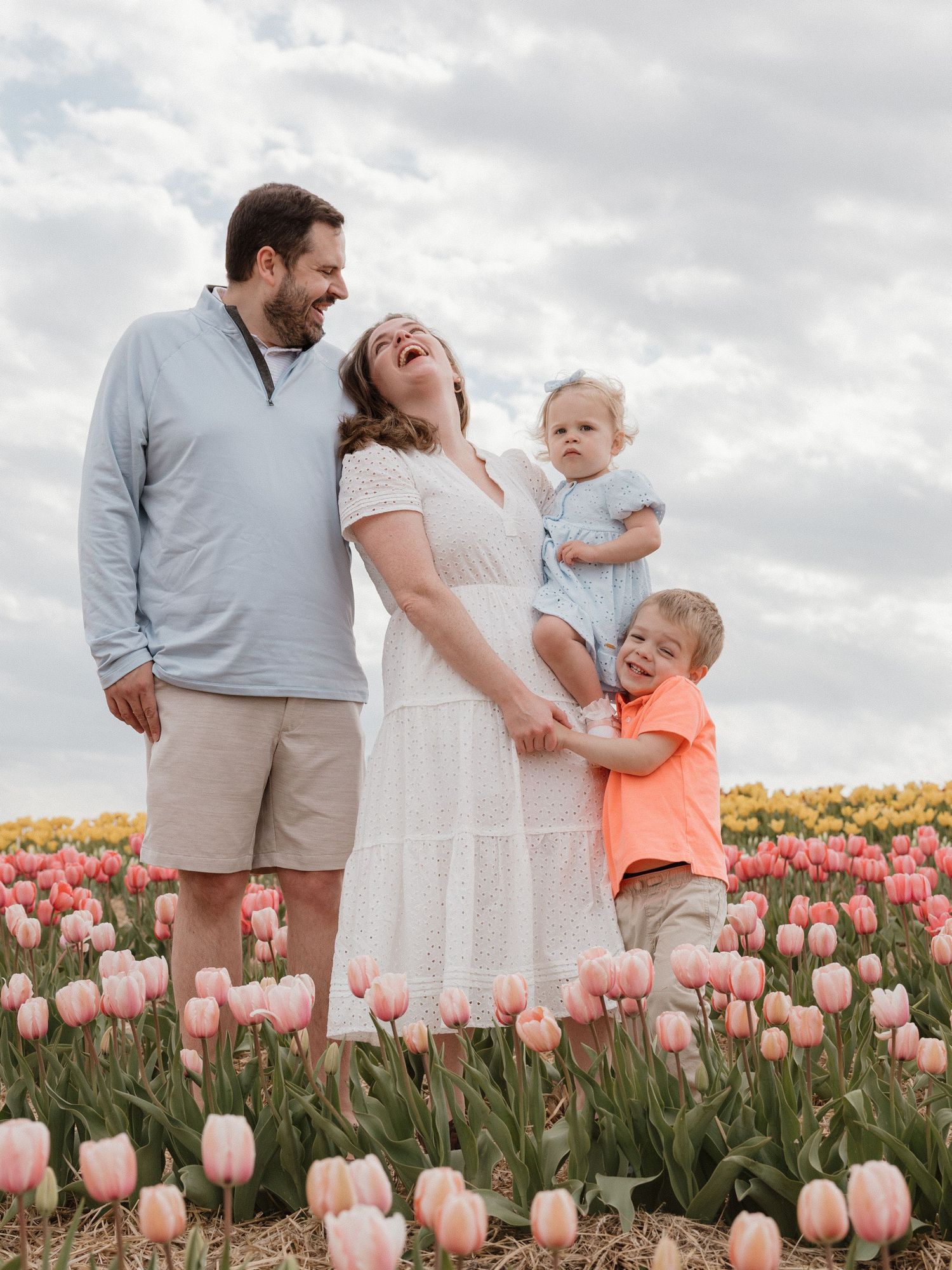 Family of four laughing together in a pink tulip field, mom holding baby in blue romper, toddler in orange shirt.