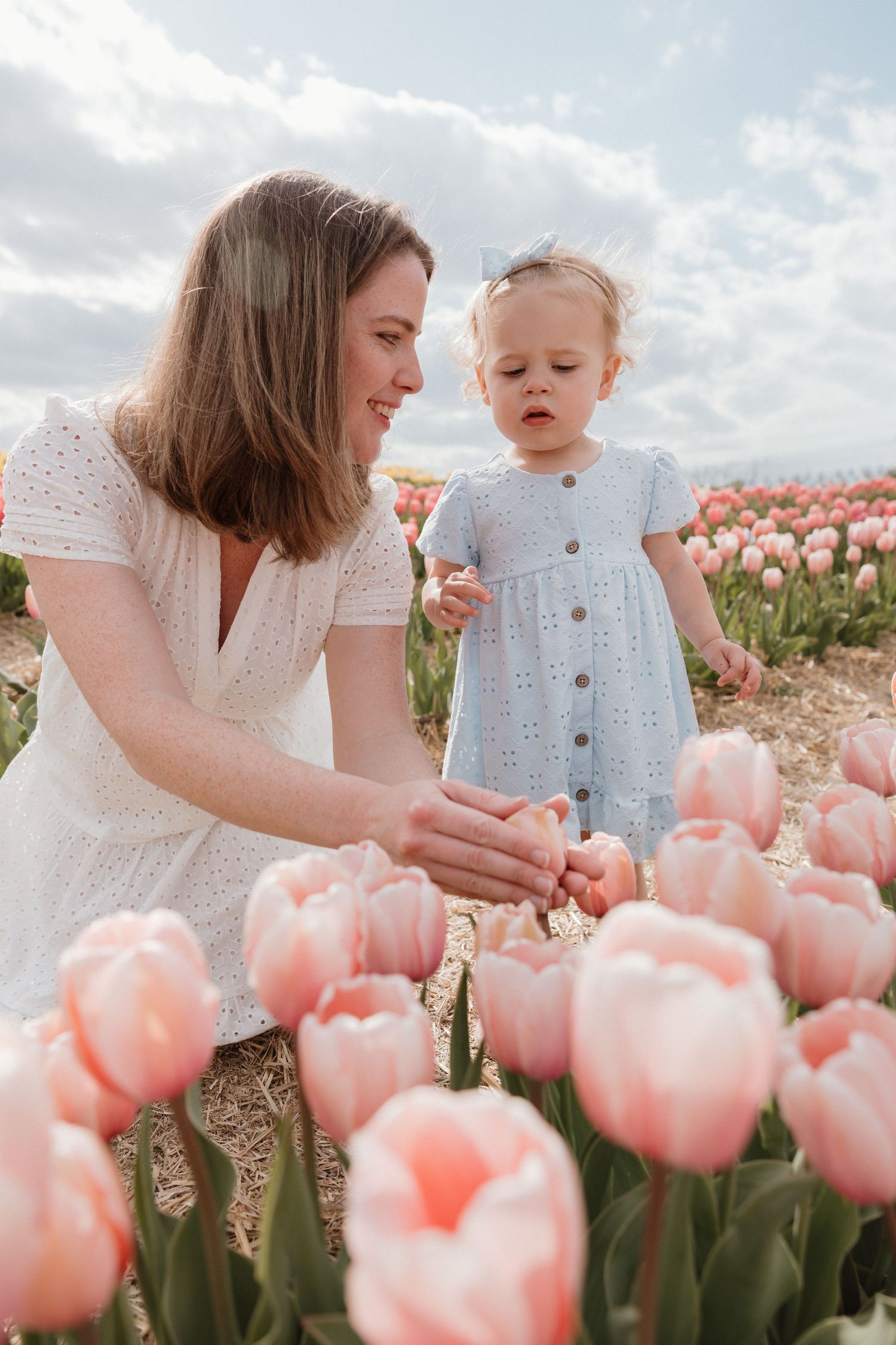 A mother and toddler girl in matching white and blue dresses explore pink tulips in a blooming flower field.
