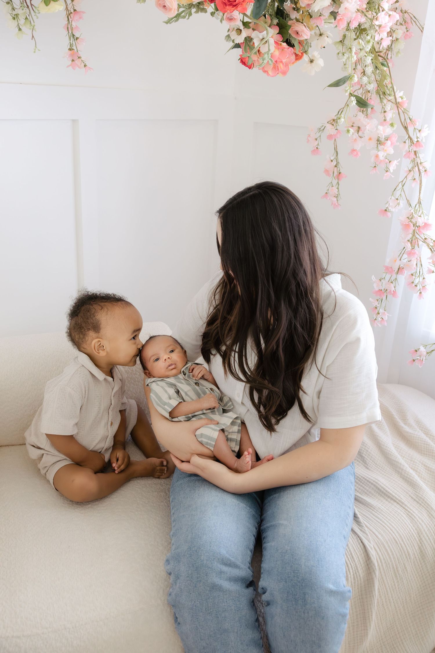 A mother in a white top and jeans holds her newborn while her toddler son gives the baby a sweet kiss.