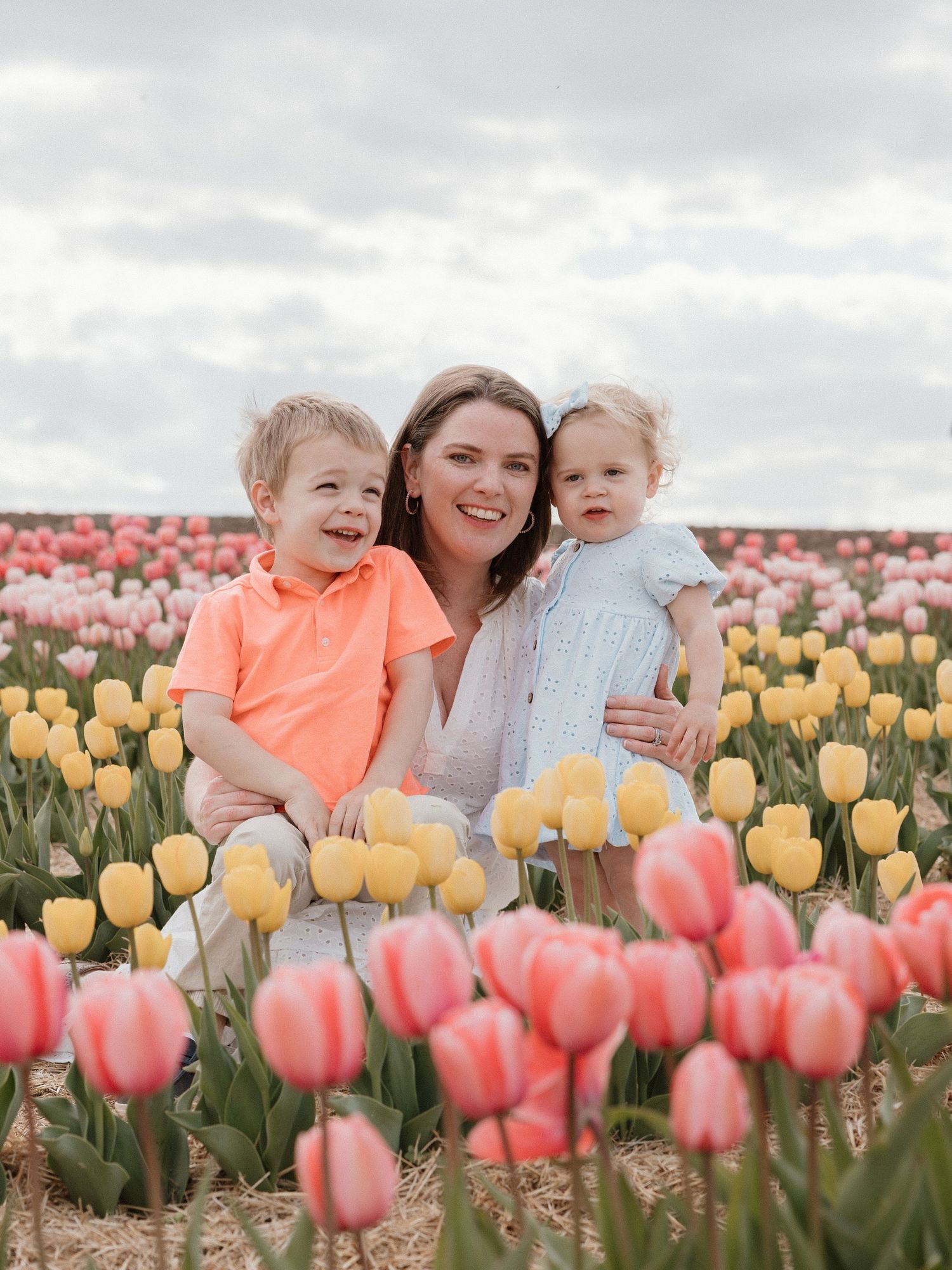 A smiling mother sits with two young children in a vibrant tulip field with pink and yellow blooms.