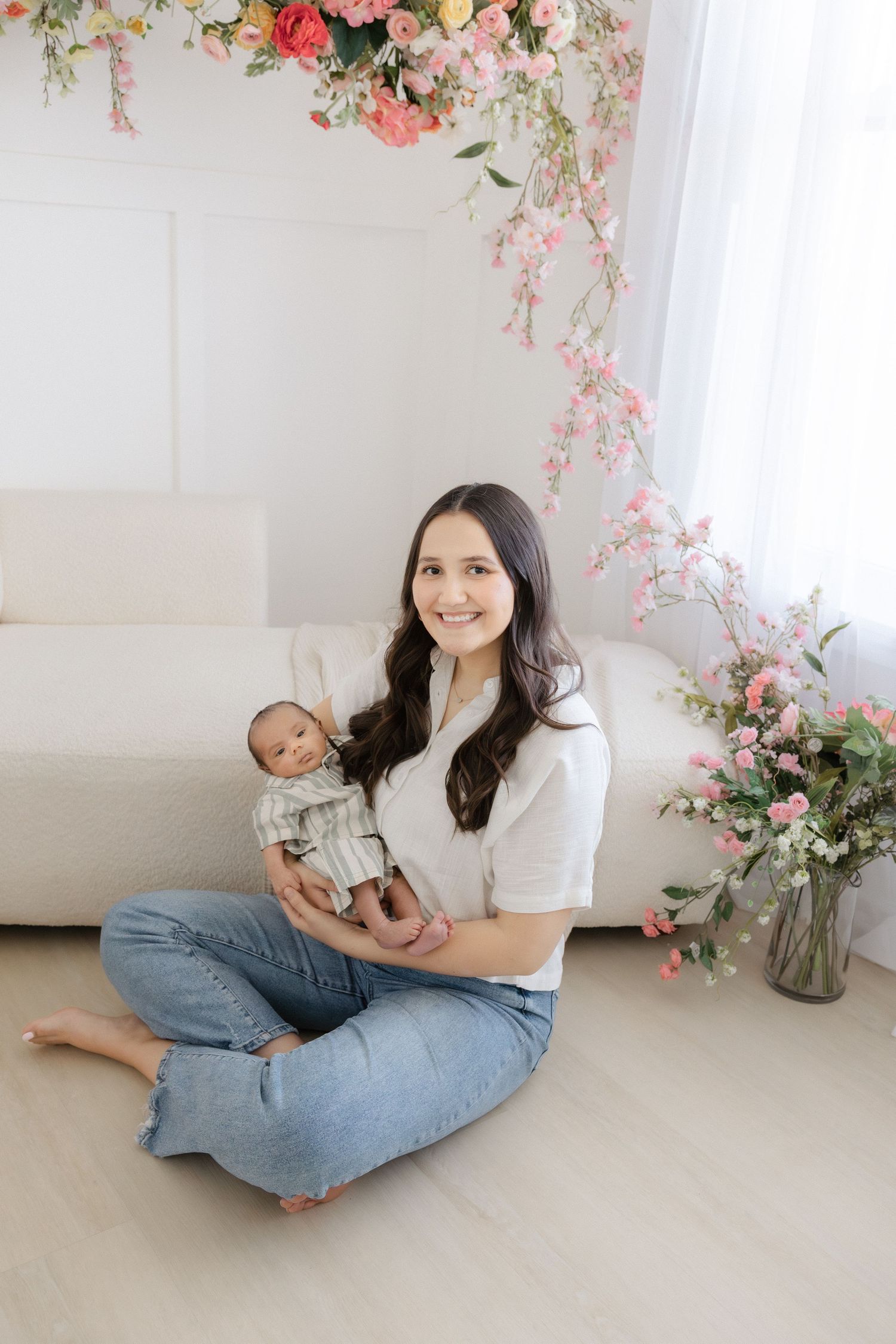 A smiling woman sits cross-legged on a white floor holding a newborn baby, surrounded by pink floral decorations.