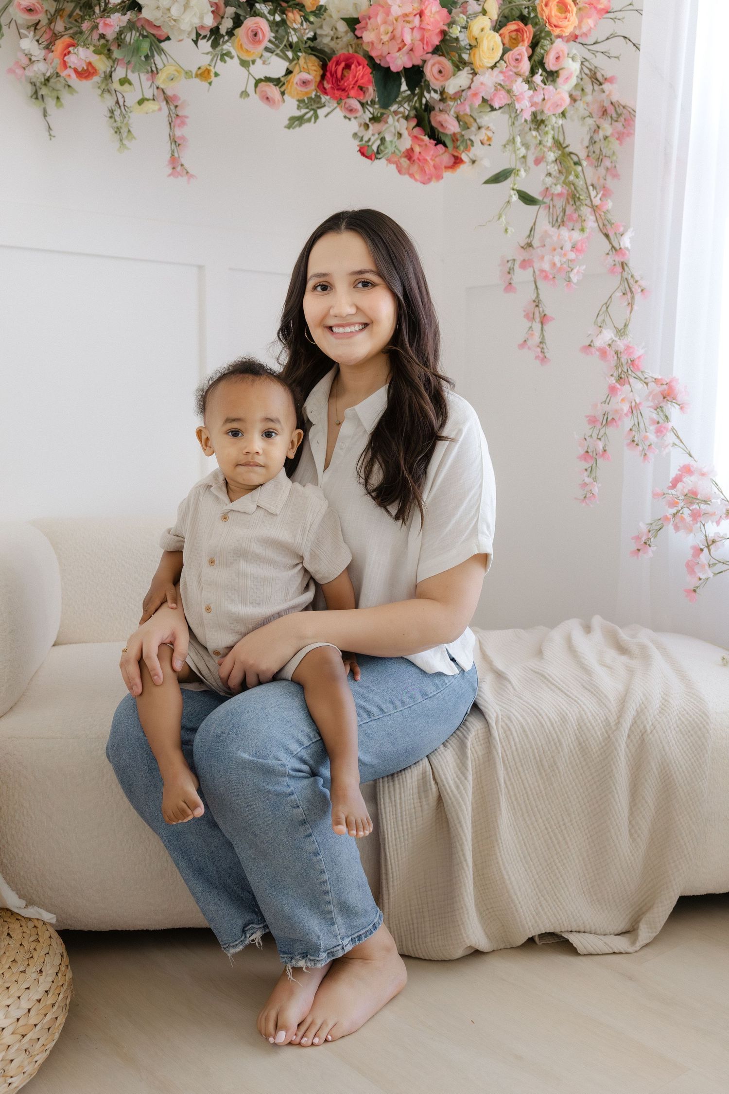 A smiling woman sits on a cream sofa holding a toddler, surrounded by pink floral decor in a bright studio.