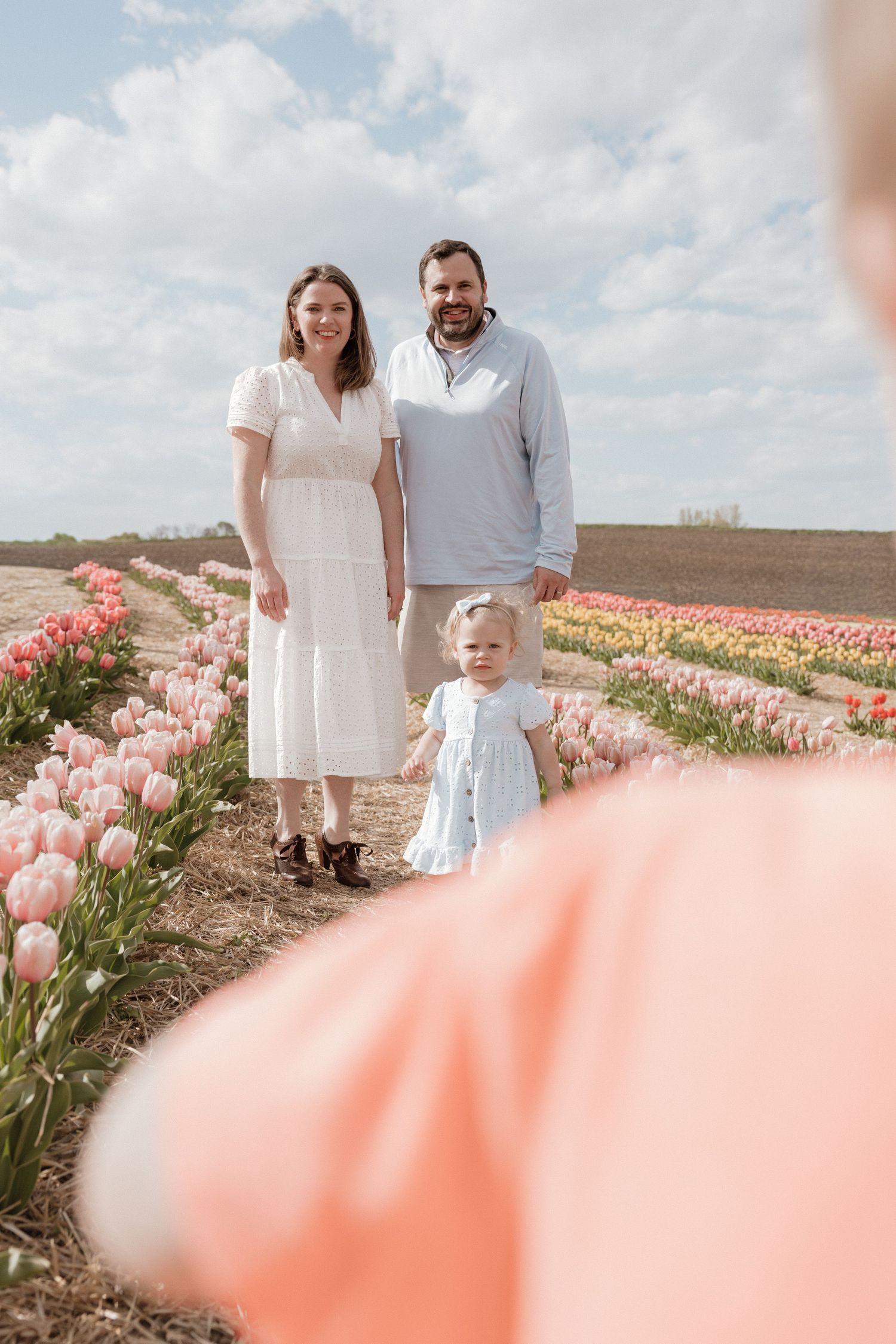 Family of three posing in a colorful tulip field on a sunny day, with a blurred foreground figure.