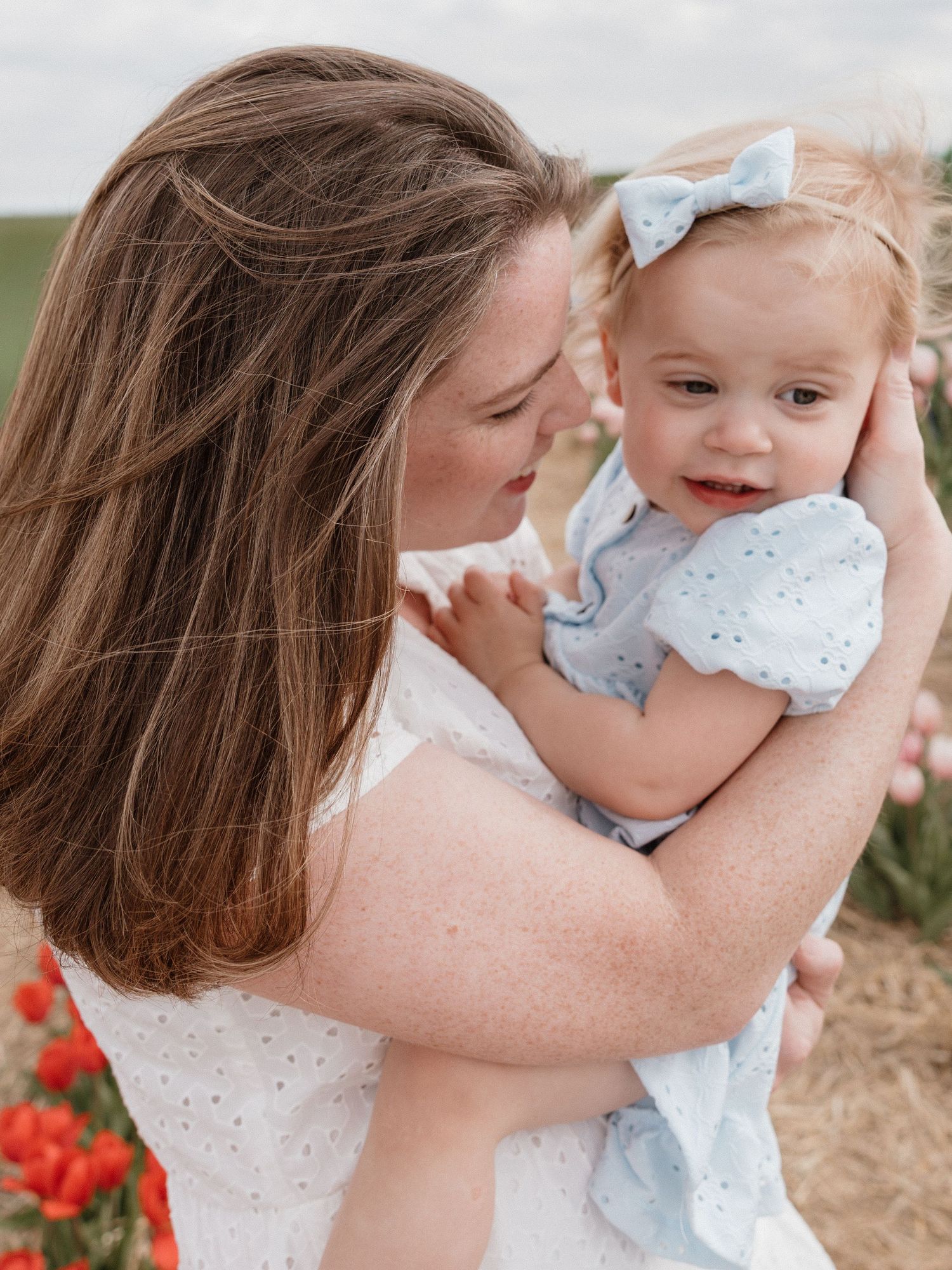 A mother in a white dress lovingly holds her smiling toddler girl in a blue dress with a bow headband outdoors.