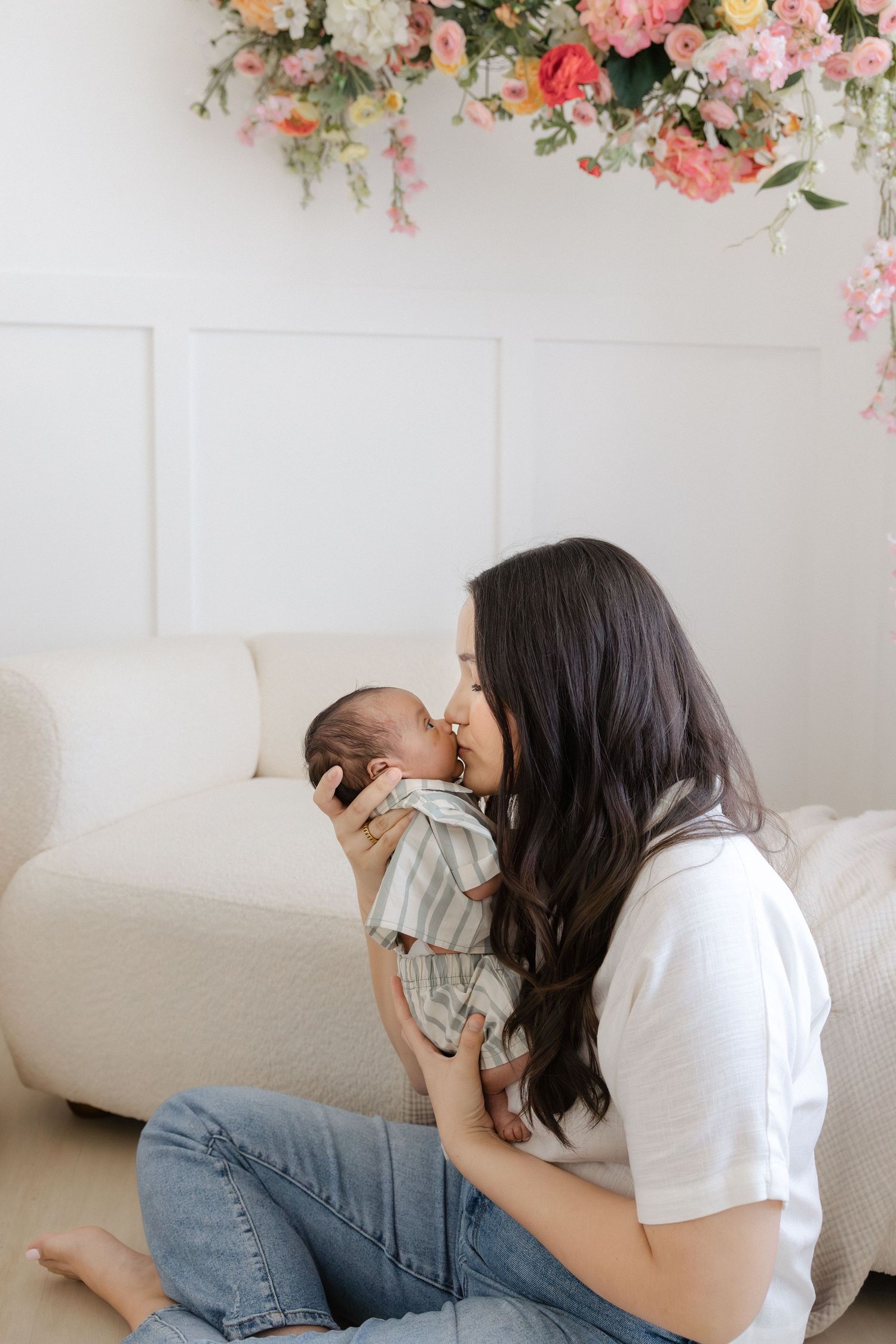 A mother gently kisses her newborn baby while sitting on a white floor, with floral decor overhead.