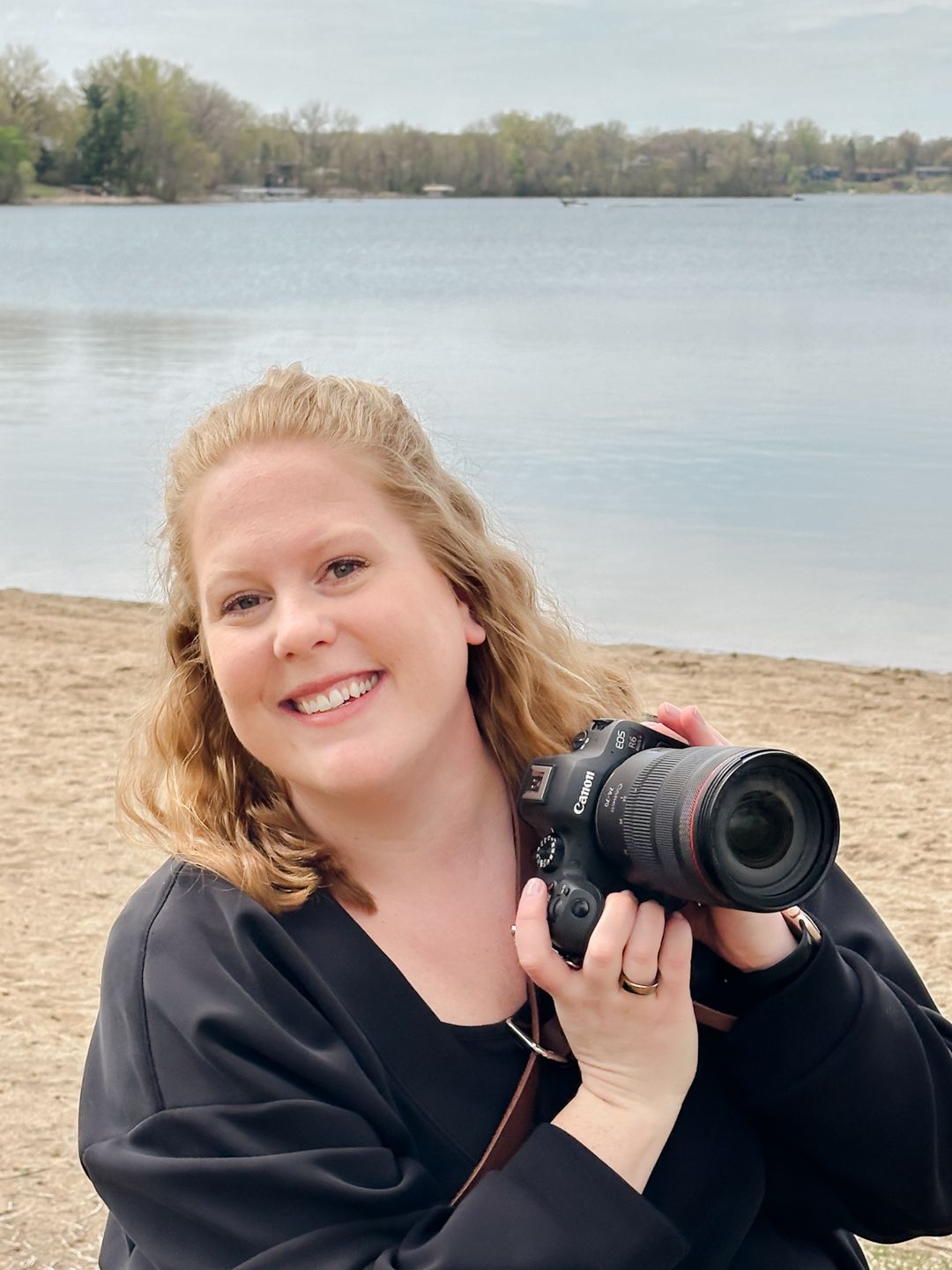 Smiling woman holding a DSLR camera near a lake shore with trees in the background on a cloudy day.