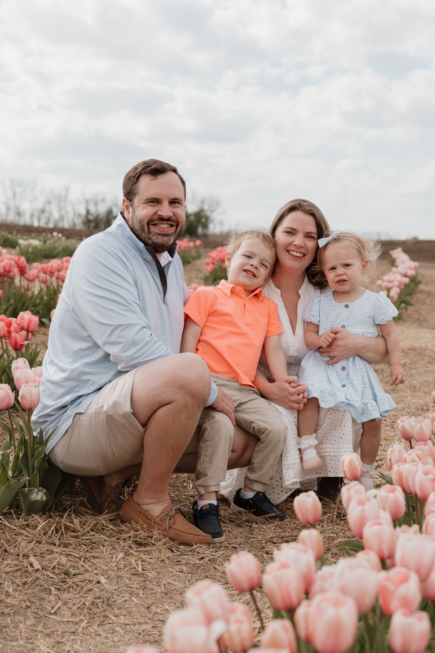 Family of four smiling in a pink tulip field, parents with young boy and toddler girl on a cloudy spring day.